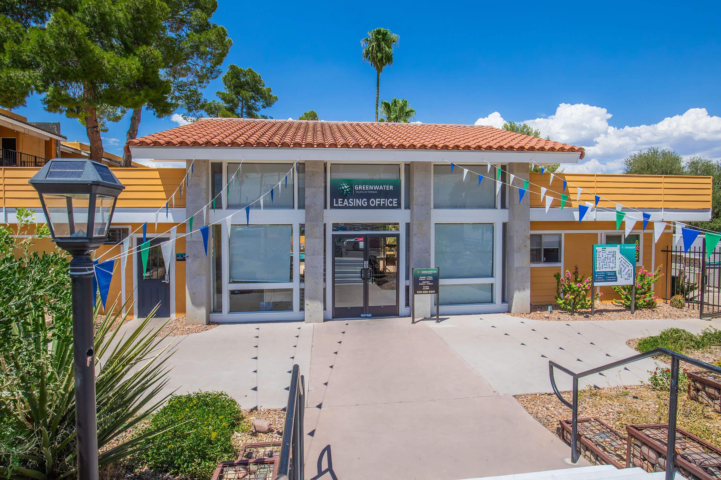 A modern leasing office with a large entrance, surrounded by desert landscaping. The building features a tile roof and is accented by colorful banners. Palm trees are visible in the background under a bright blue sky with scattered clouds. A lamp post is located in the foreground, enhancing the inviting atmosphere.