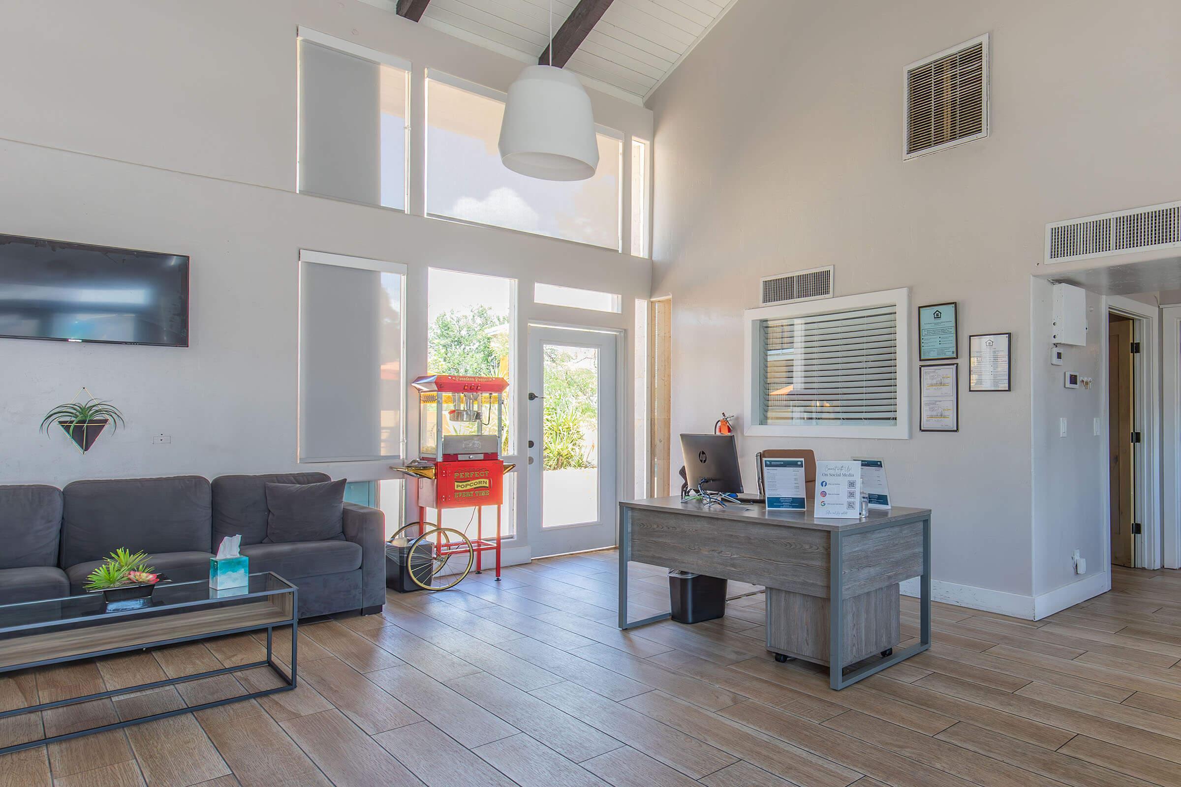 A modern reception area featuring a gray sofa, a wooden desk with a computer, and a television mounted on the wall. Large windows allow natural light, and a popcorn machine is visible in the background. The decor is simple and welcoming, with light wood flooring and neutral-colored walls.