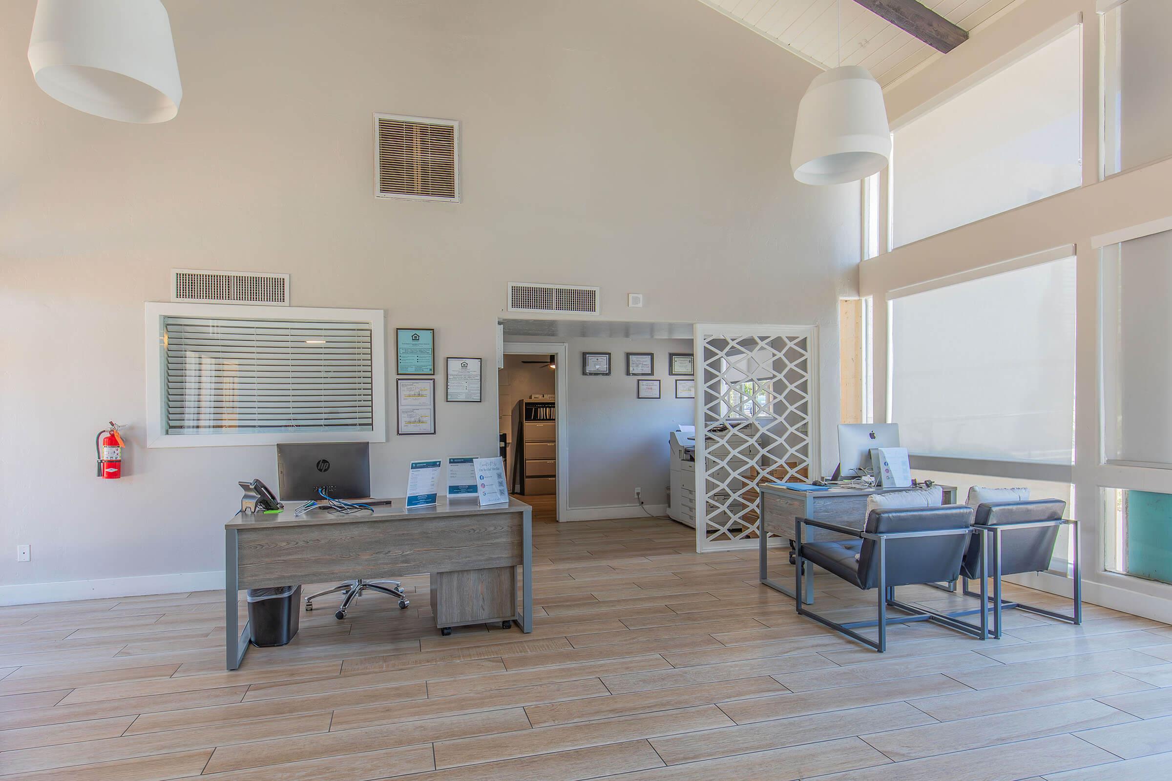 Interior of a modern office reception area. Features a large window allowing natural light, a front desk with a computer and paperwork, and a seating area with a table and chairs. The decor includes simple wall art and neutral colors, creating a welcoming and professional atmosphere.