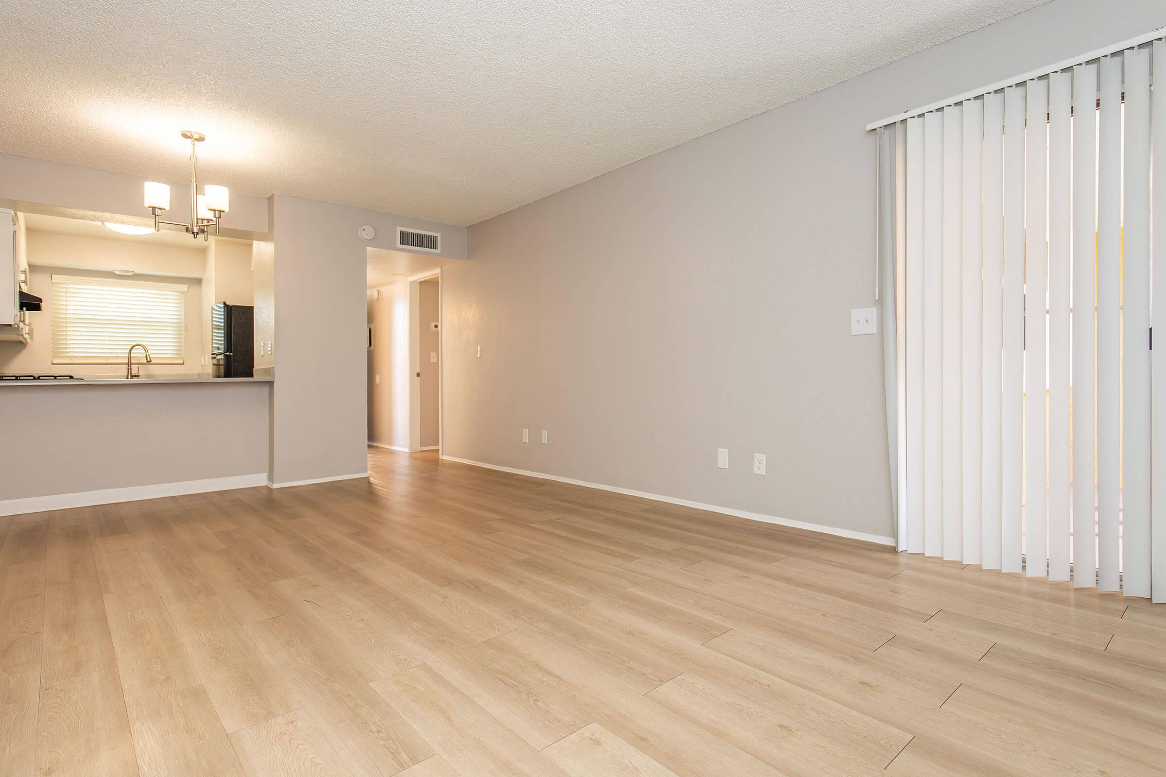 A bright and spacious living area featuring light-colored flooring, neutral walls, and a ceiling light fixture. On one side, there is a kitchen area visible with cabinets and a window, while a doorway leads to another room. Vertical blinds cover the window, creating a modern and inviting atmosphere.
