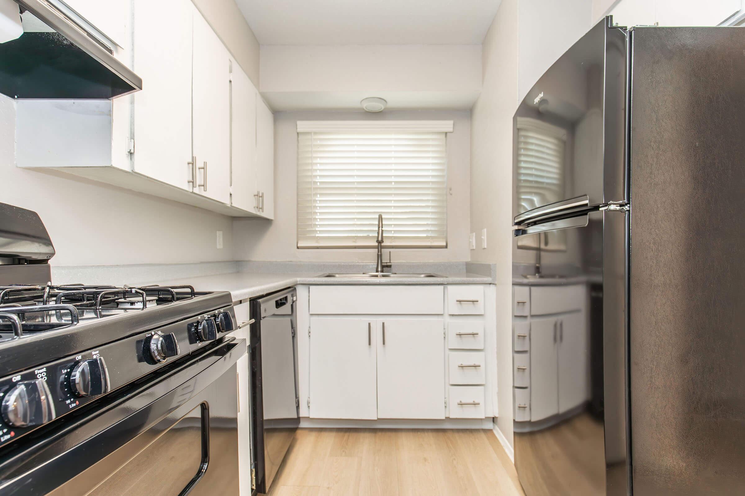 Modern kitchen featuring white cabinetry, a stainless steel refrigerator, a gas stove, and a sink under a window with blinds. The countertops are light gray, and the flooring is wood-like laminate. The design is clean and contemporary, suitable for small living spaces.
