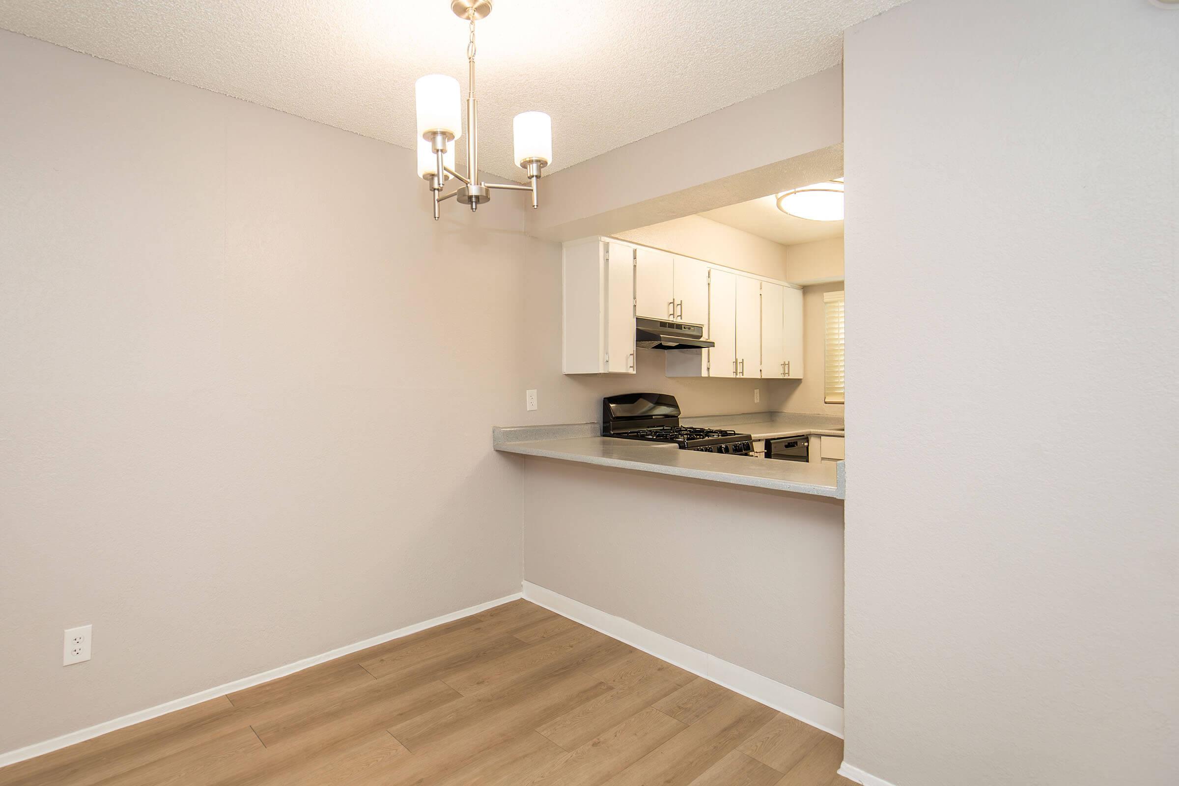 Modern kitchen area featuring a light-colored wall and wood flooring. The kitchen has white cabinets and a grey countertop, with a gas stove and oven. A small opening connects the kitchen to the living space. Ceiling lights provide illumination in the room.