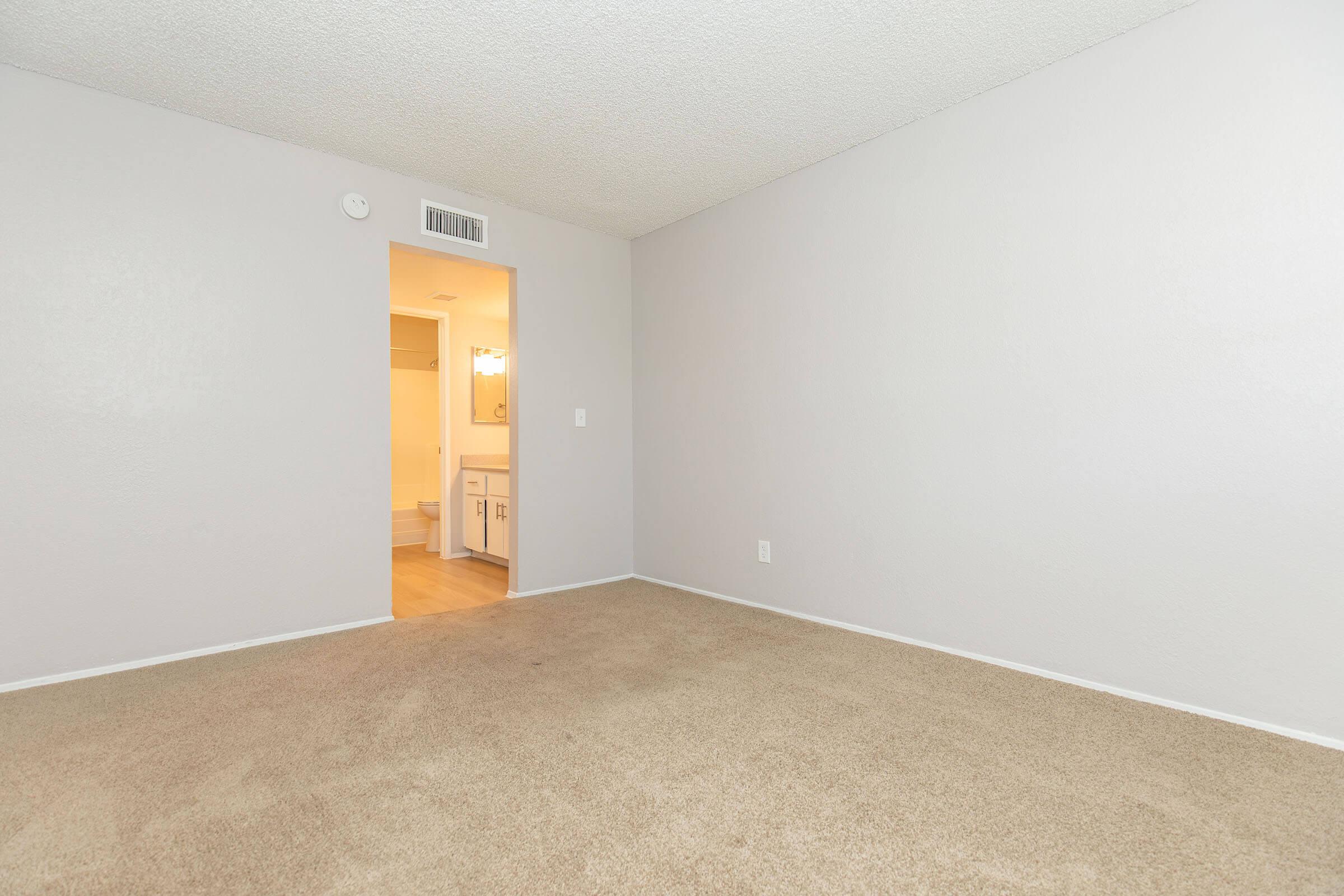 A spacious, empty room with light gray walls and beige carpeting. There is a doorway leading to a well-lit bathroom area, featuring a light fixture visible in the background. The room is unoccupied, conveying a clean and neutral atmosphere, suitable for various uses or personal decoration.