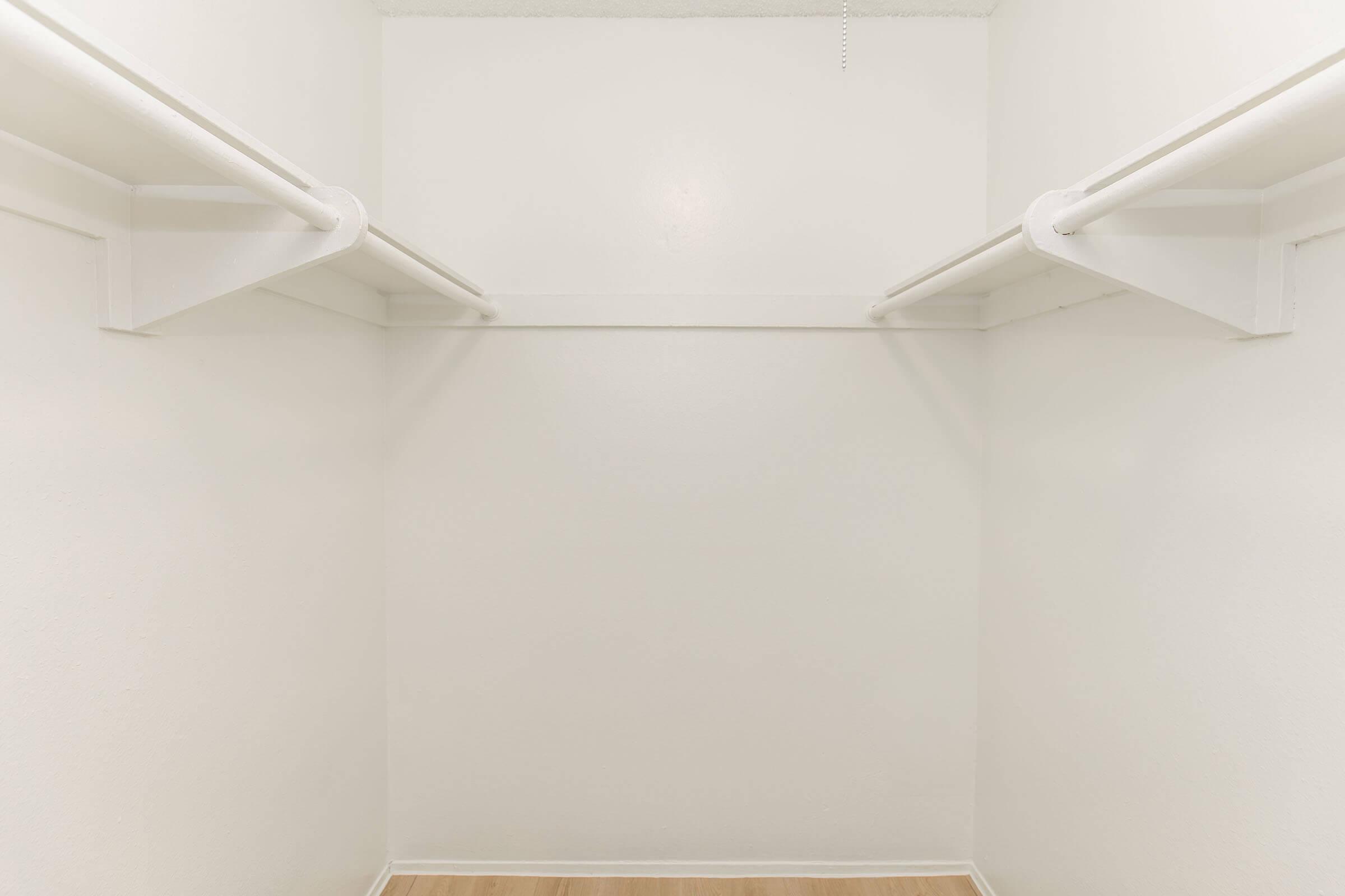 A spacious, empty closet with white walls and two hanging rods along the sides. The floor is light-colored wood, creating a clean and minimalistic appearance. The room is well-lit, emphasizing the uncluttered space.