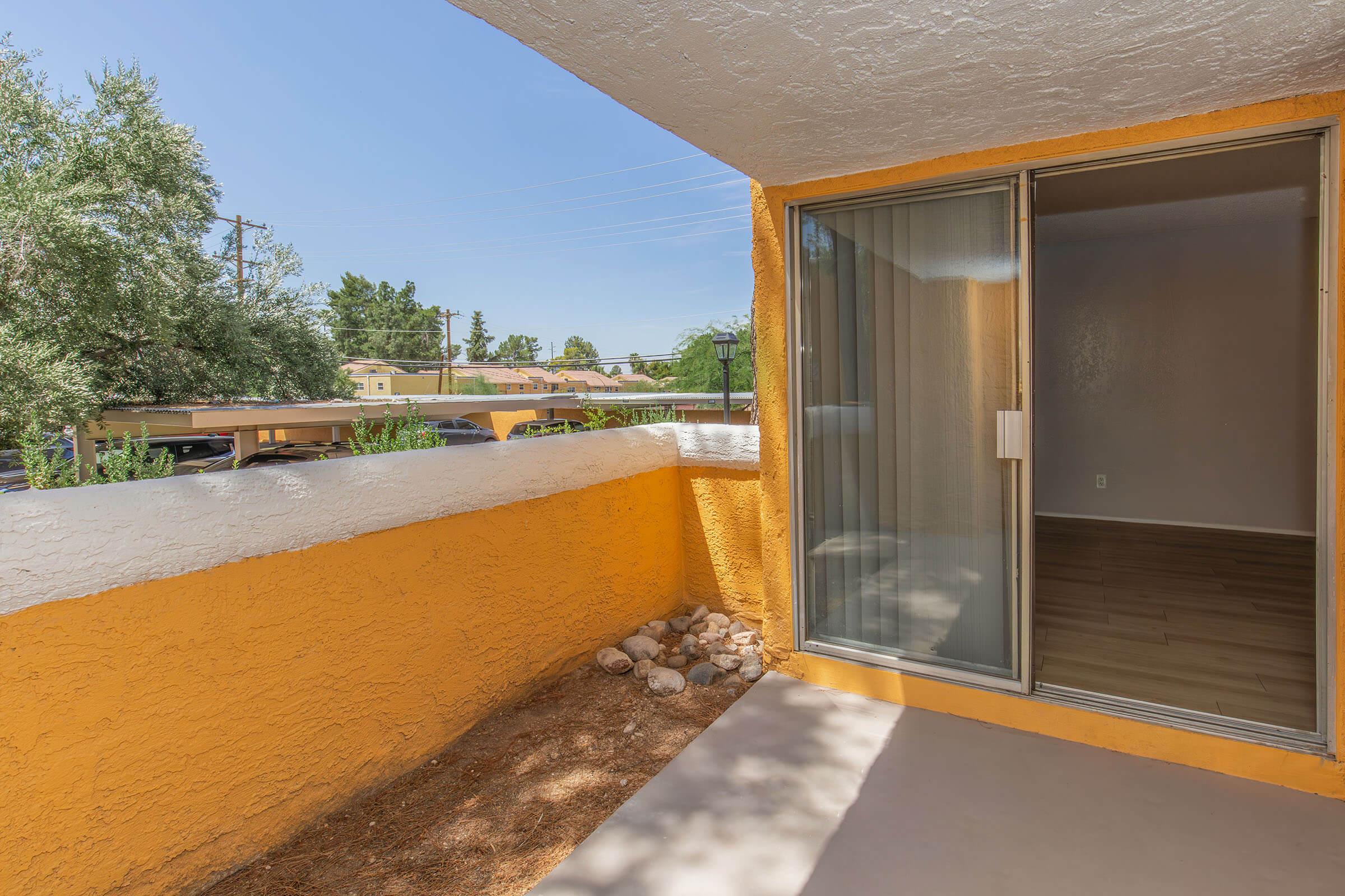 A small balcony with an orange textured wall and a sliding glass door. The floor is light-colored, and there are some rocks and gravel on the ground. In the background, trees and parked vehicles are visible under a clear blue sky.