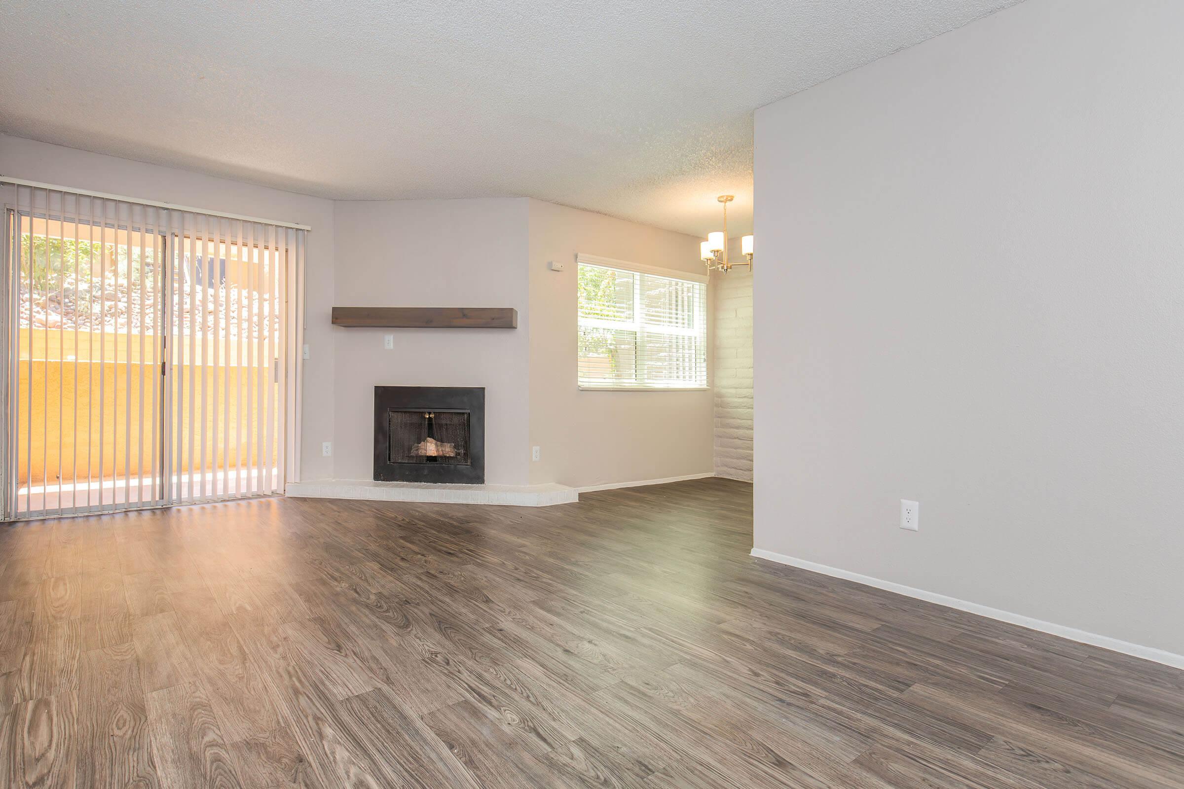 Spacious living area featuring laminate flooring, a decorative fireplace, and large windows allowing natural light. A sliding door leads to a balcony, and a dining area is visible with a chandelier. The walls are painted in light neutral tones.