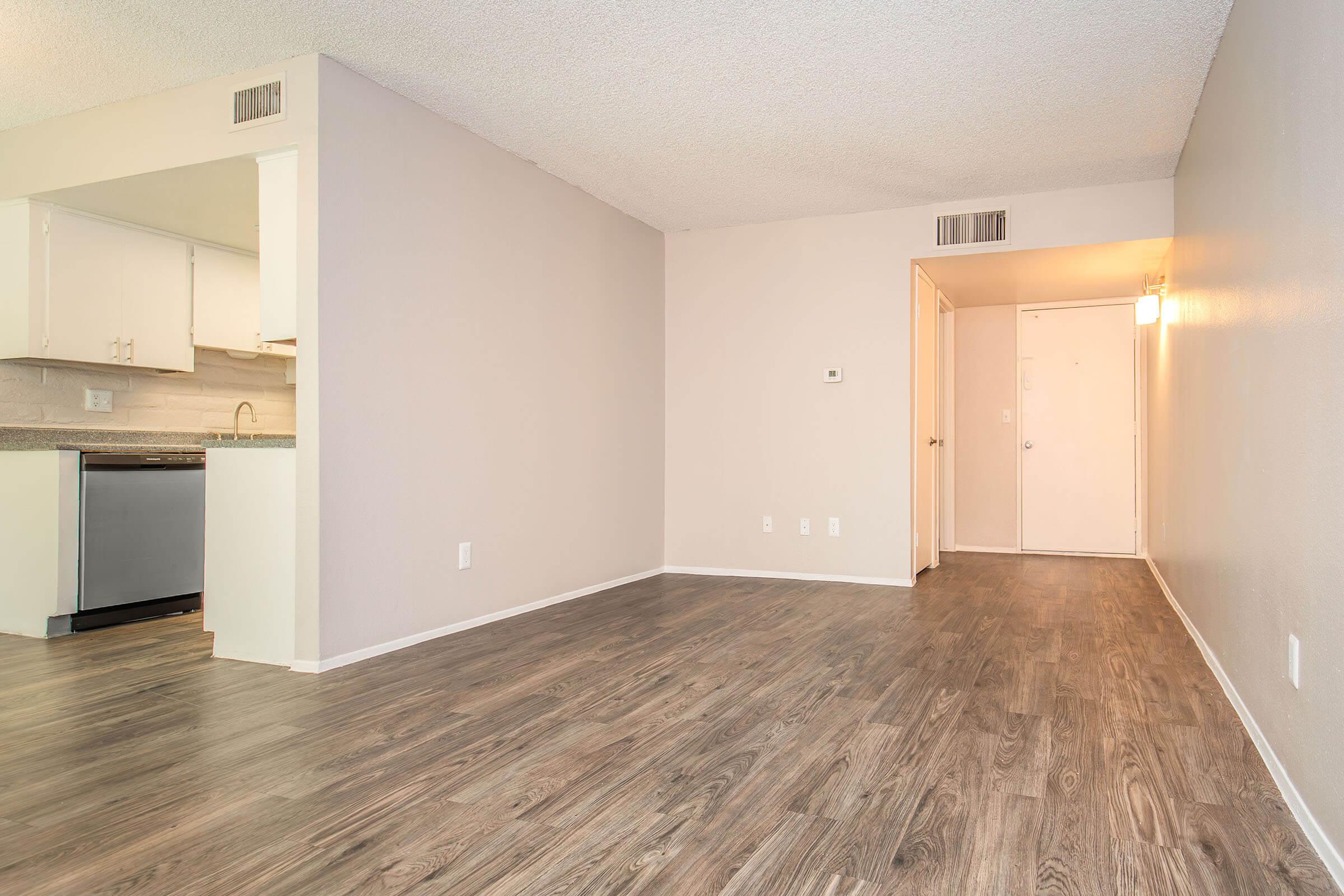 Spacious interior of an apartment showcasing a clean, neutral-colored living area with hardwood flooring. The layout includes a doorway leading to a hallway and a glimpse of a kitchen area with modern appliances. Ideal for showcasing a bright, open floor plan.