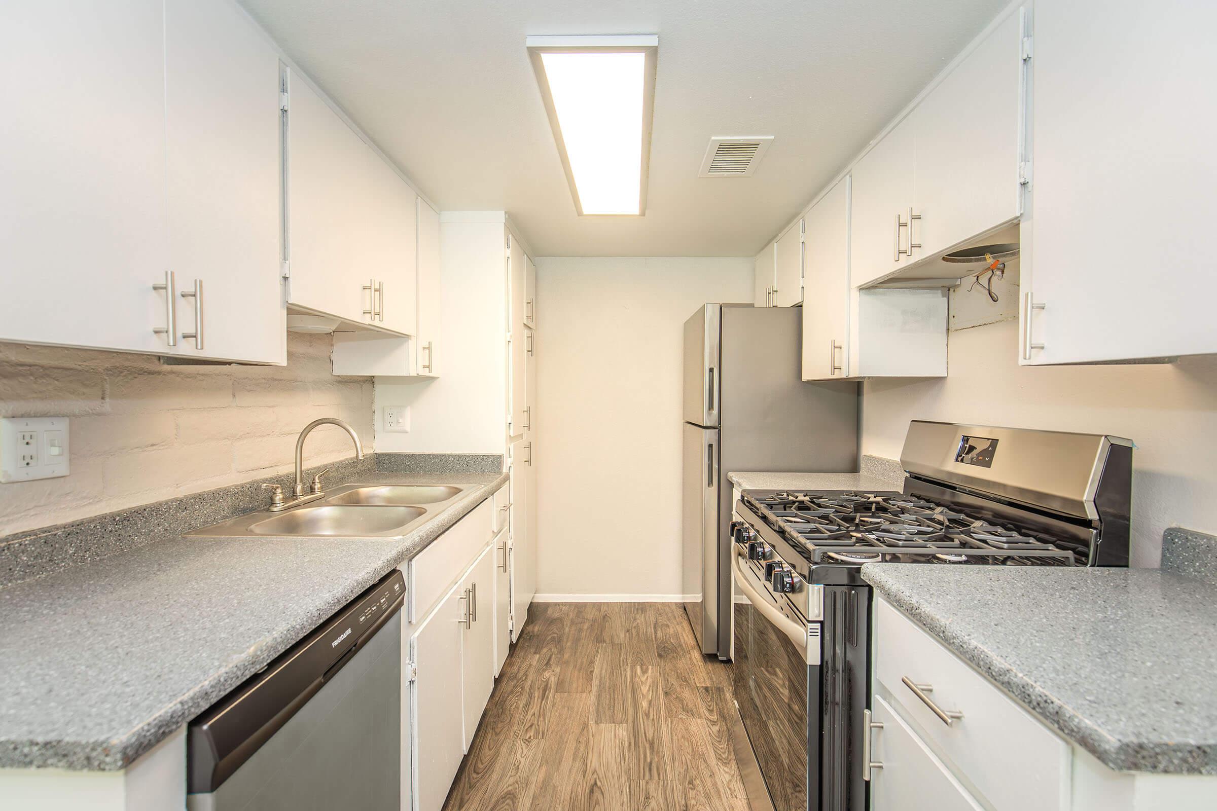 A modern kitchen featuring white cabinets, stainless steel appliances including a refrigerator and gas stove, a double sink, and granite countertops. The space has a bright overhead light and a clean, minimalist design with wood-style flooring.