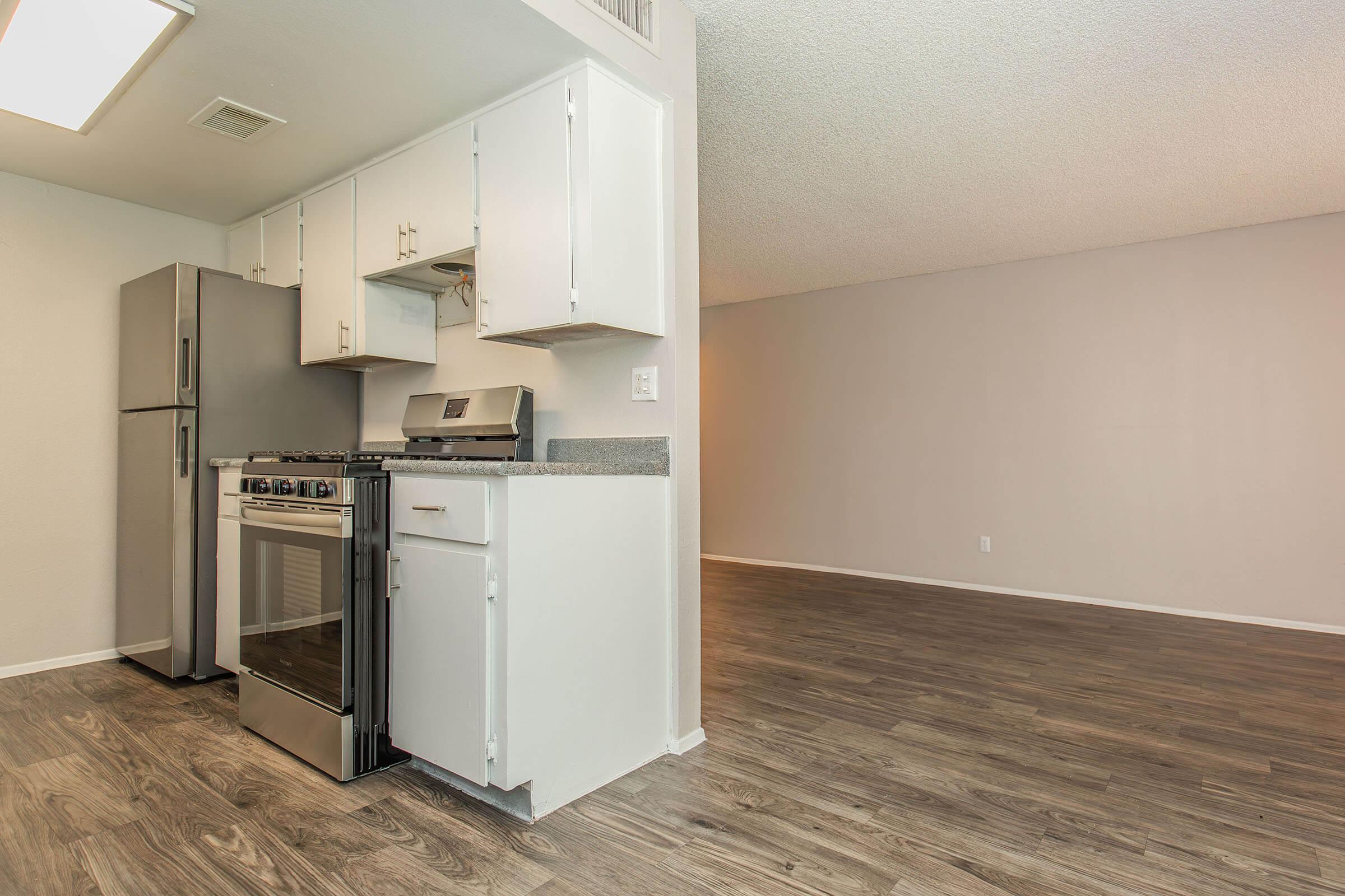 A modern kitchen with stainless steel appliances, including a refrigerator and oven, situated next to a spacious living area. The kitchen features white cabinets and granite countertops, with a light-colored wall and wood-like flooring throughout. Natural light fills the space, creating an inviting atmosphere.