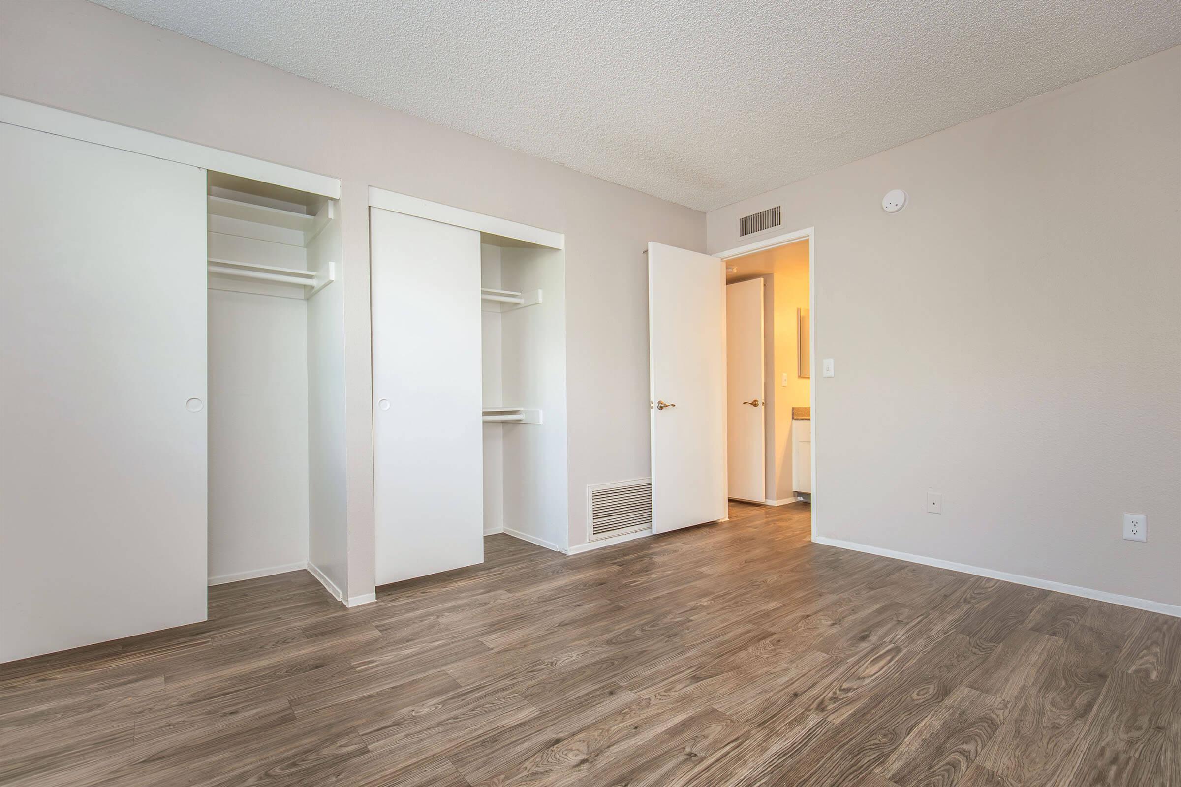 A spacious room featuring light-colored walls and wood-like flooring. Two built-in white closets are on the left, and an open doorway to a bathroom or hallway can be seen in the background on the right. Natural light brightens the area, creating a fresh and inviting atmosphere.
