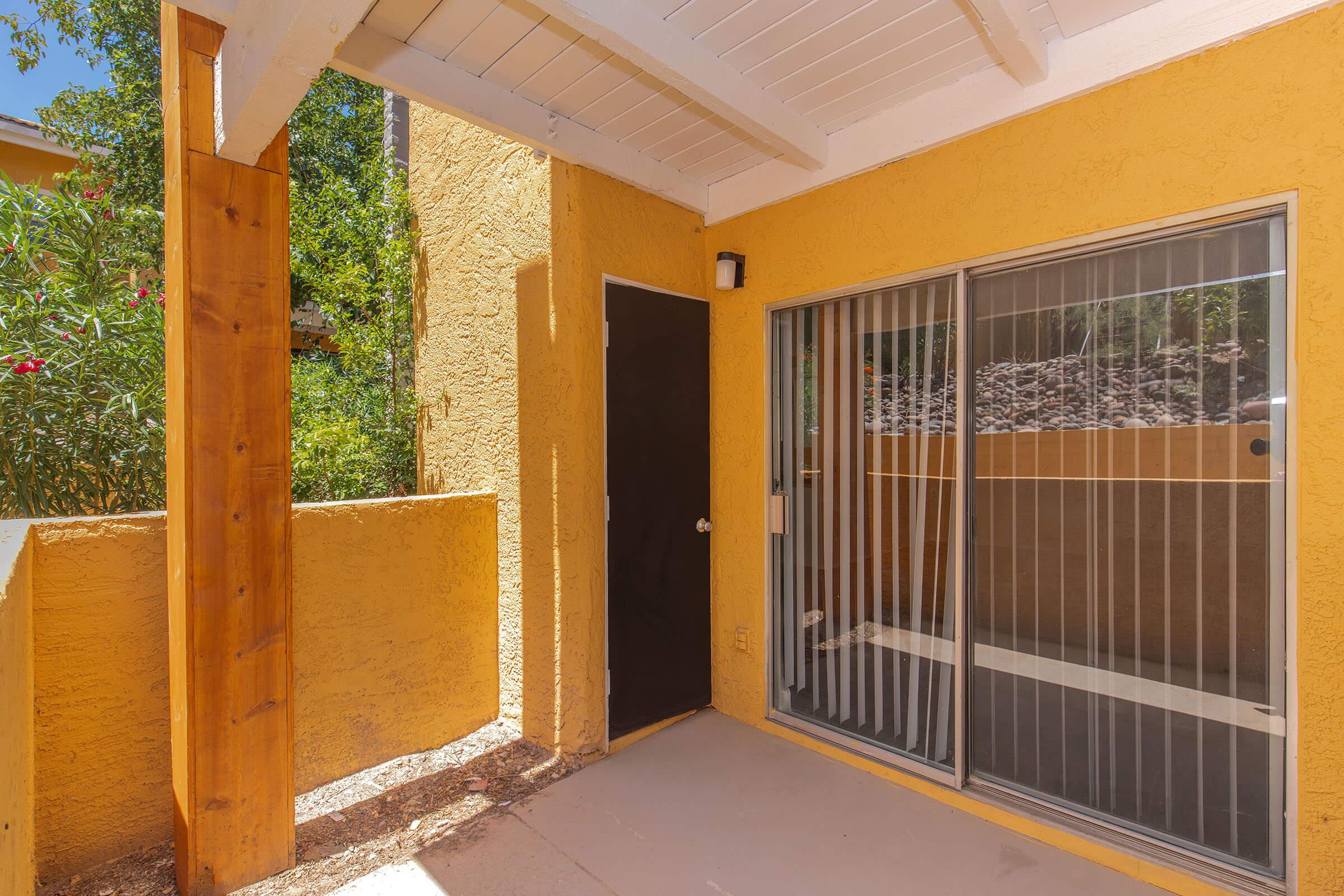 A view of a yellow exterior wall with a sliding glass door, surrounded by greenery. The door is framed by wooden pillars, and there is a small patio area in front. The space is bright and inviting, with sunlight casting shadows on the ground.