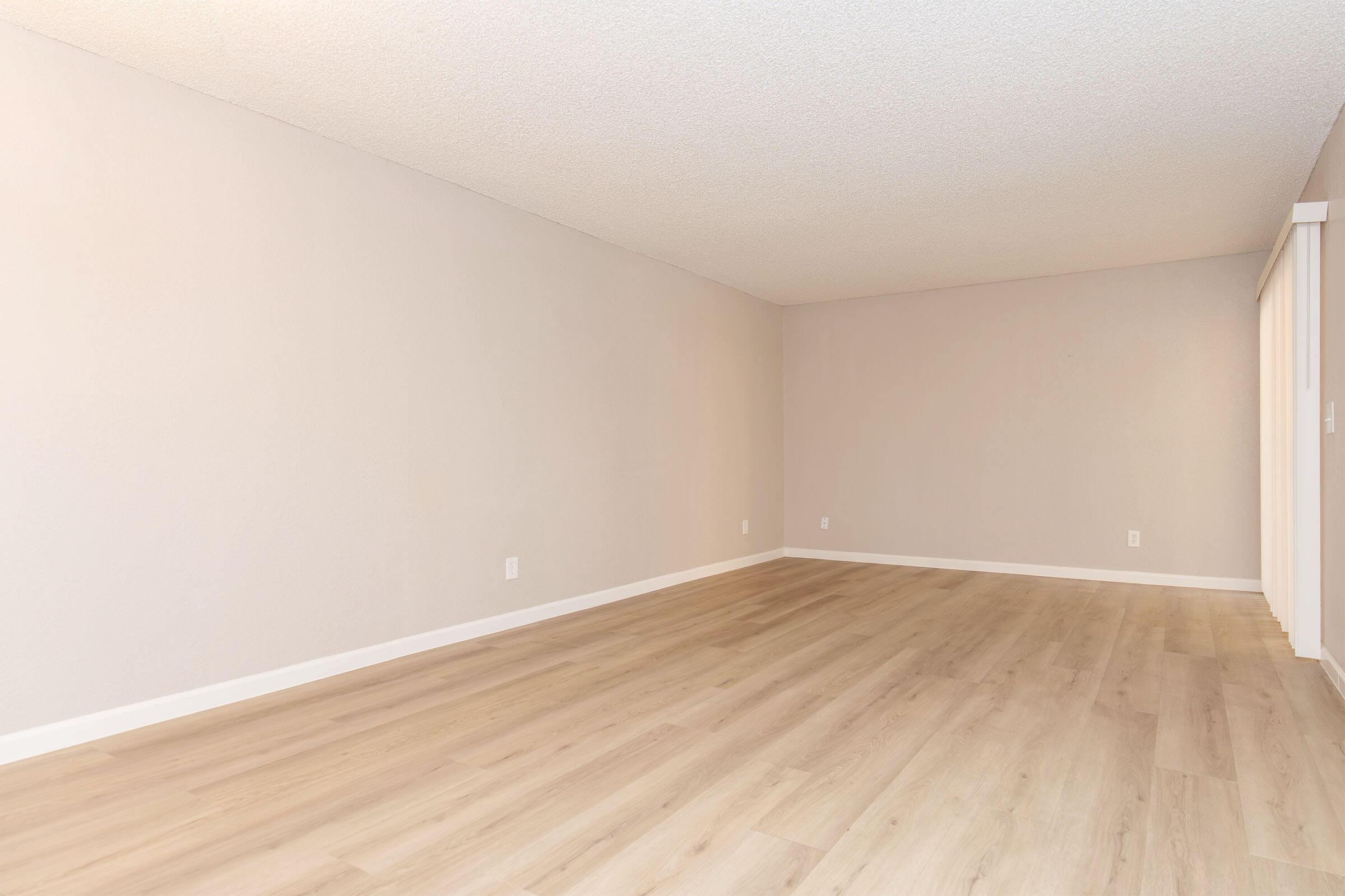 An empty room with light-colored walls and a smooth wooden floor. There are no furniture or decorations in the space, creating a minimalistic and open atmosphere. The ceiling features a textured surface, and a corner shows a partial wall with sliding doors.