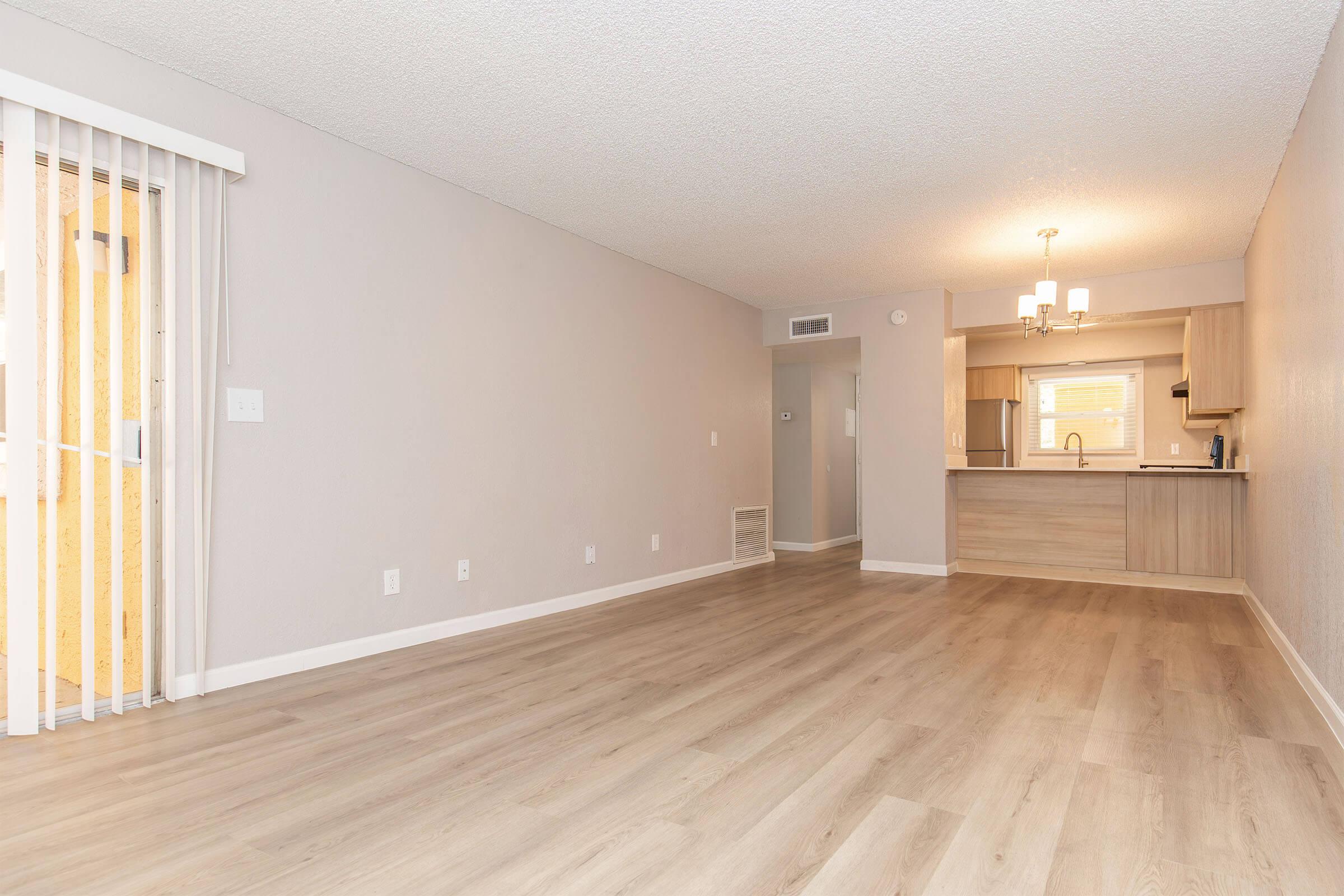 Spacious and modern living room with light wood flooring, neutral-colored walls, and a sliding glass door leading to an exterior space. The kitchen area features a breakfast bar and contemporary cabinetry, illuminated by a ceiling light fixture.