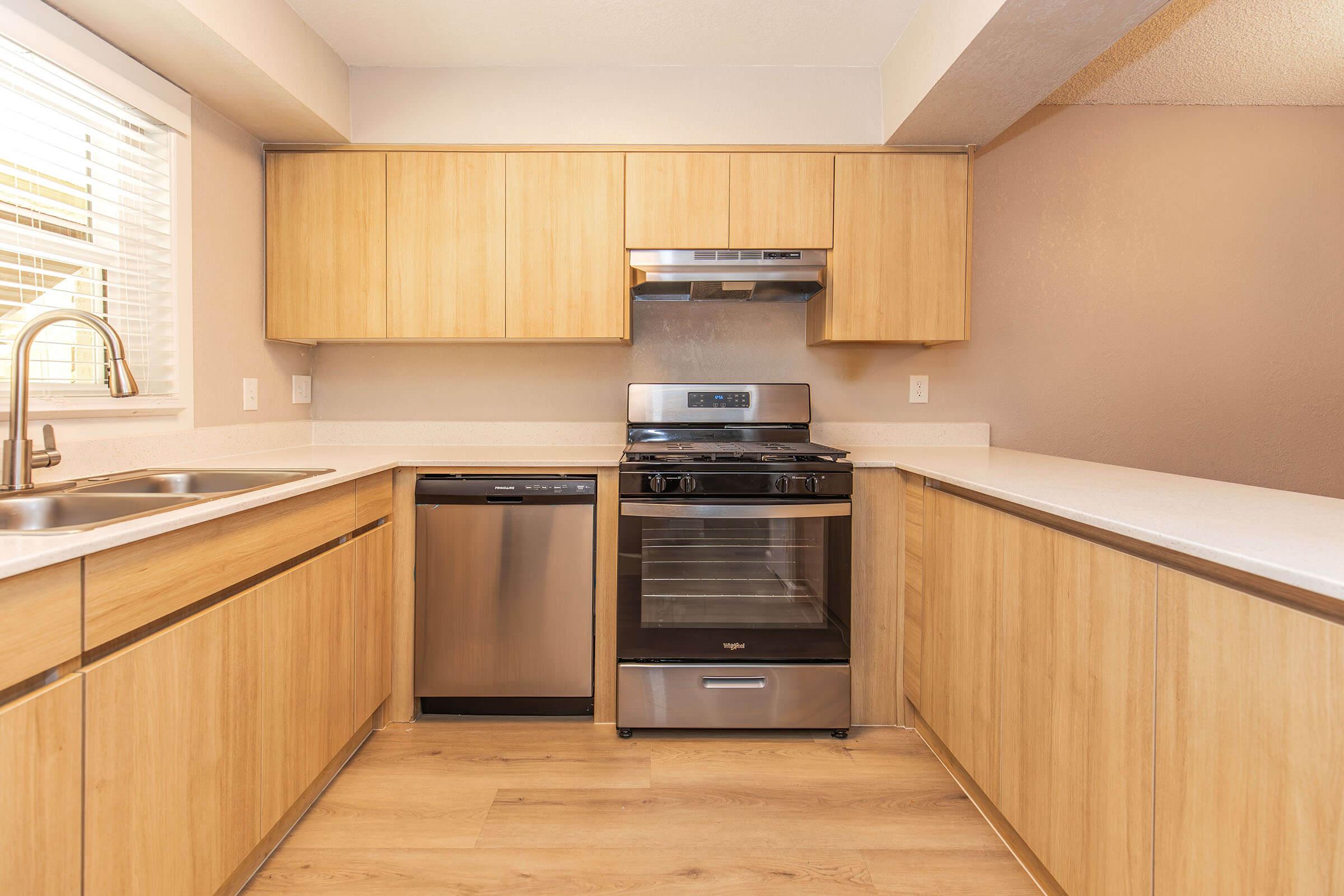 Modern kitchen featuring light wood cabinetry, stainless steel appliances including a dishwasher and a range with an overhead microwave. The countertop is a neutral tone, and the kitchen has a bright, airy feel with natural light coming through a window.