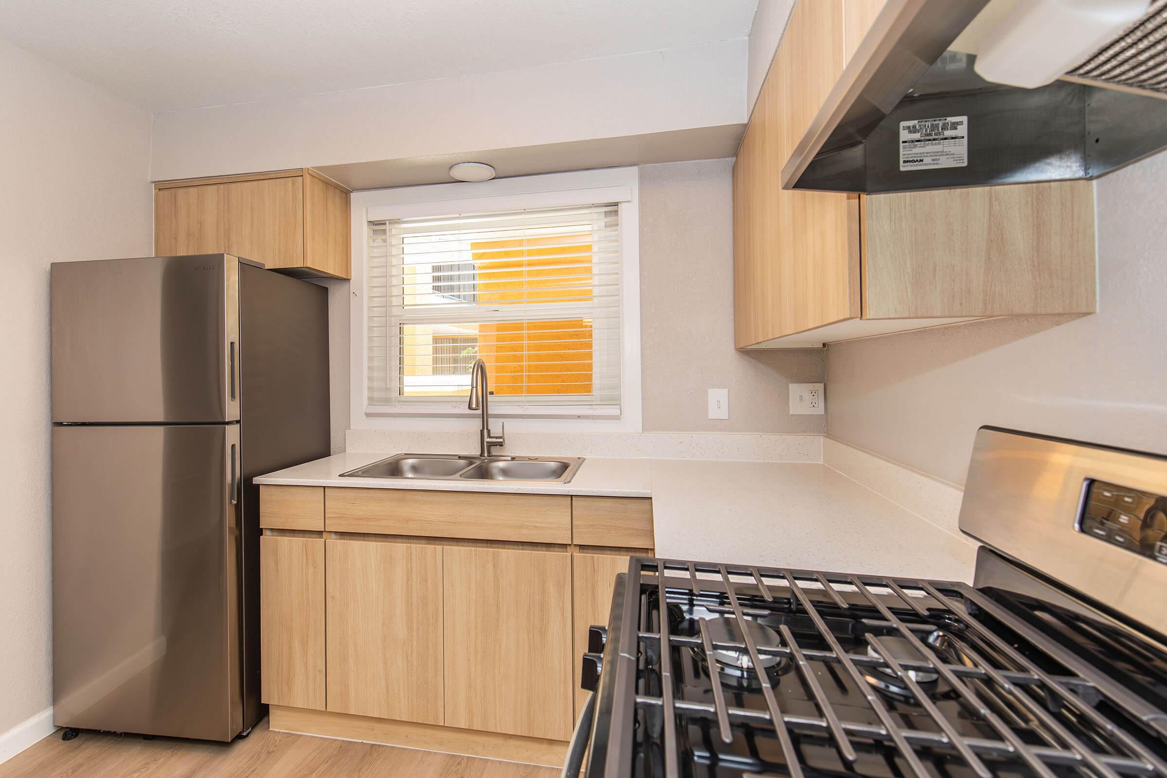Modern kitchen featuring wooden cabinetry, a stainless steel refrigerator, and a gas stove with an oven. A window above the sink allows natural light to brighten the space, which is adorned with neutral colors and wooden flooring.