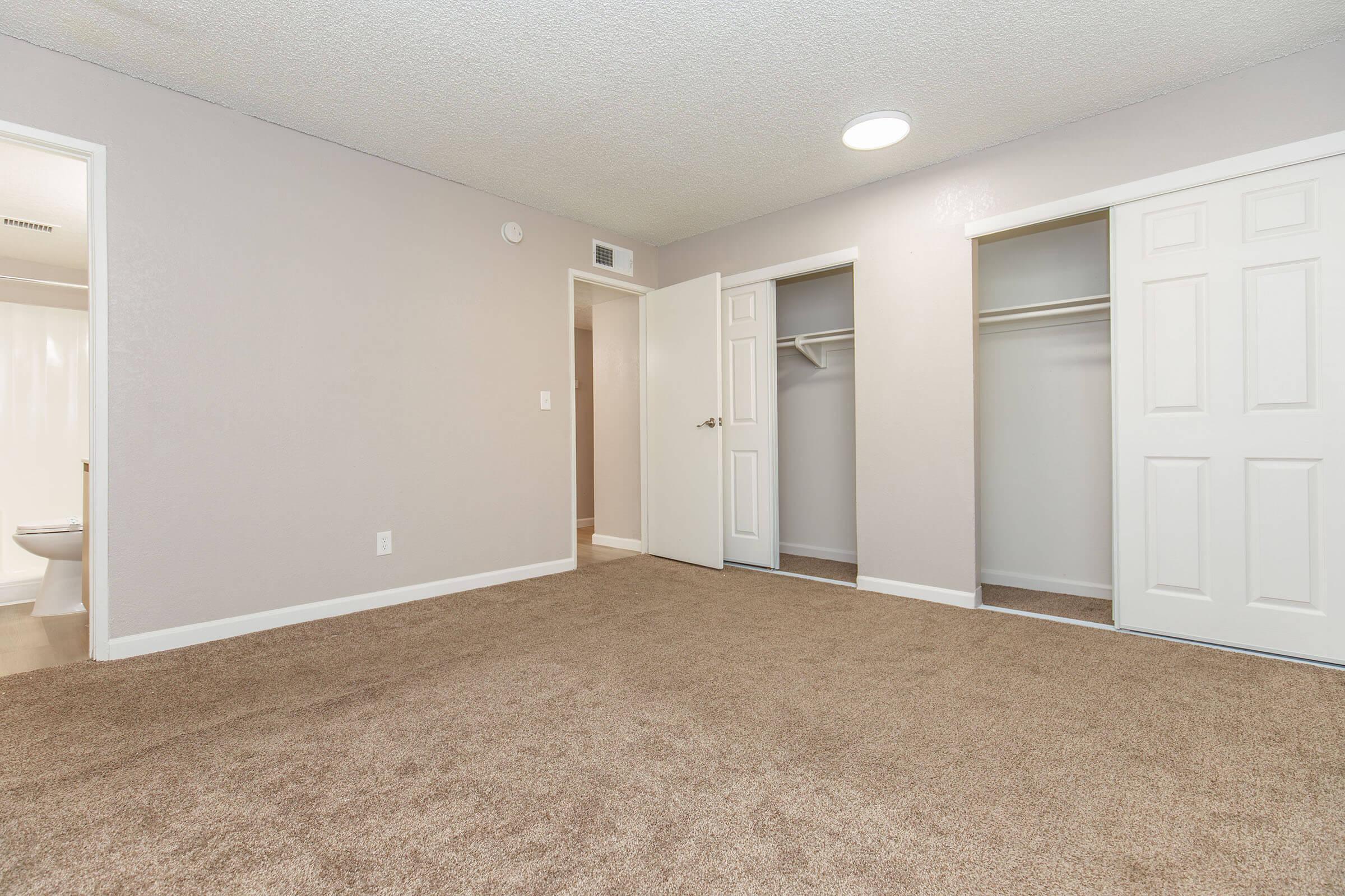 A spacious, empty bedroom with light beige carpeting and light gray walls. There are two closet doors on one side and a door leading to another room. The ceiling has a round light fixture, and a small bathroom is visible in the background through an open door. The room is well-lit and uncluttered.