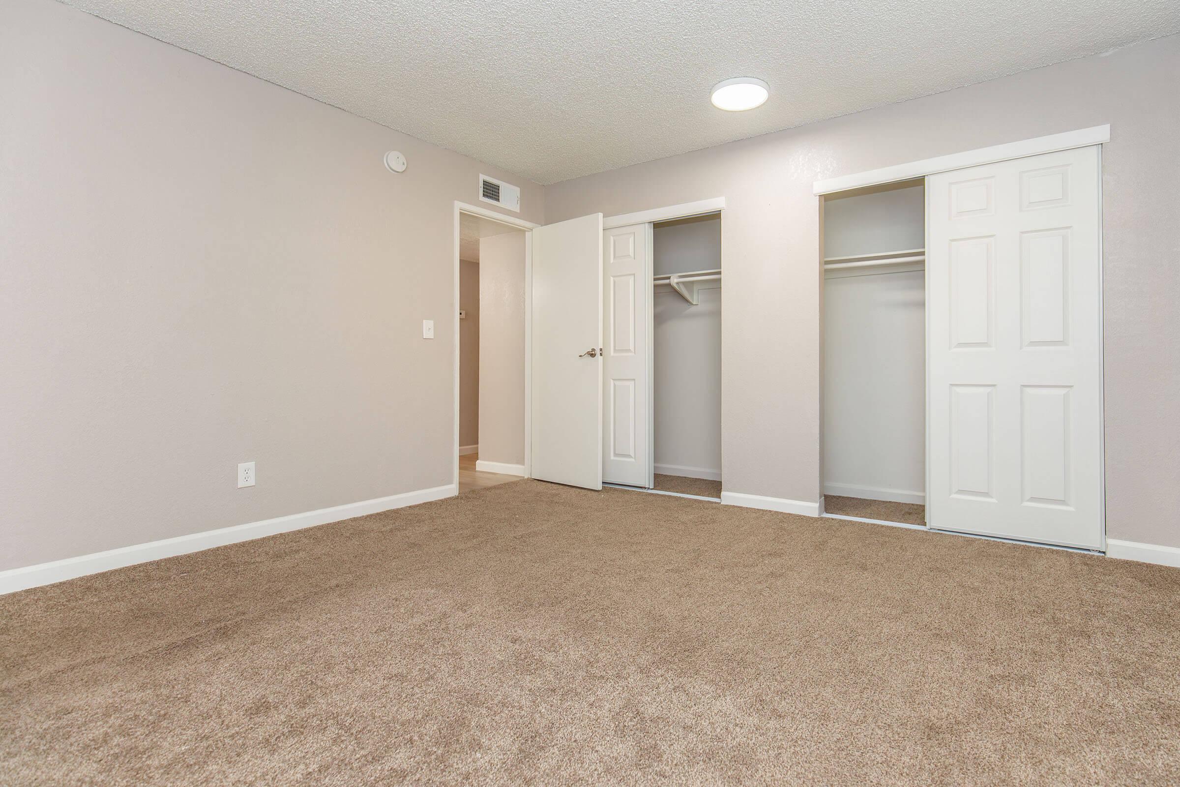 A spacious, well-lit bedroom with a neutral-colored carpet and light purple walls. Two white closet doors are visible on the right, and a doorway leads to another room on the left. The ceiling features a circular light fixture, contributing to a bright and inviting atmosphere.