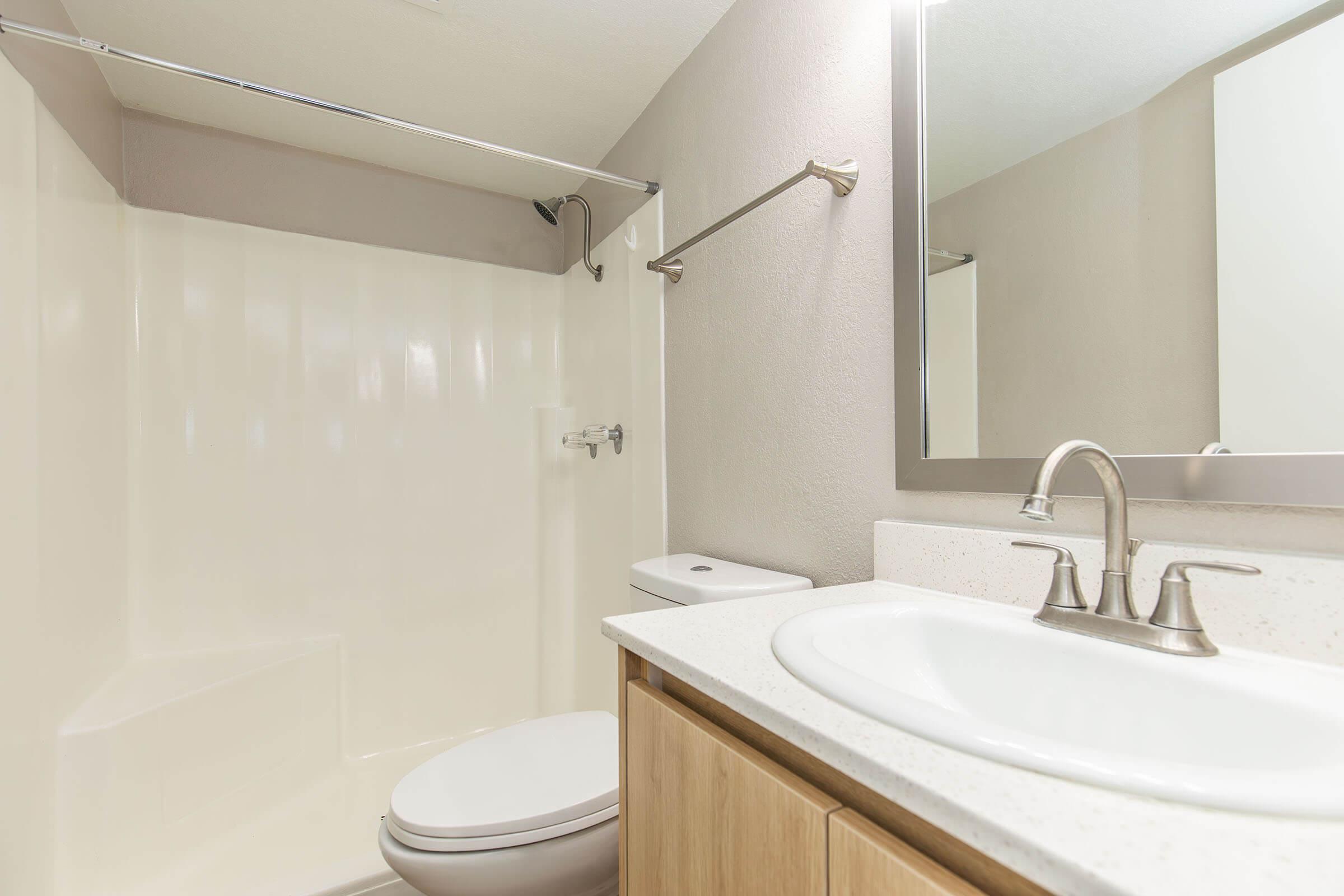 A clean and modern bathroom featuring a white shower stall, toilet, and sink with a light-colored countertop. The walls are a neutral tone, and there's a large mirror above the sink. The overall design is simple and contemporary, emphasizing functionality and minimalism.