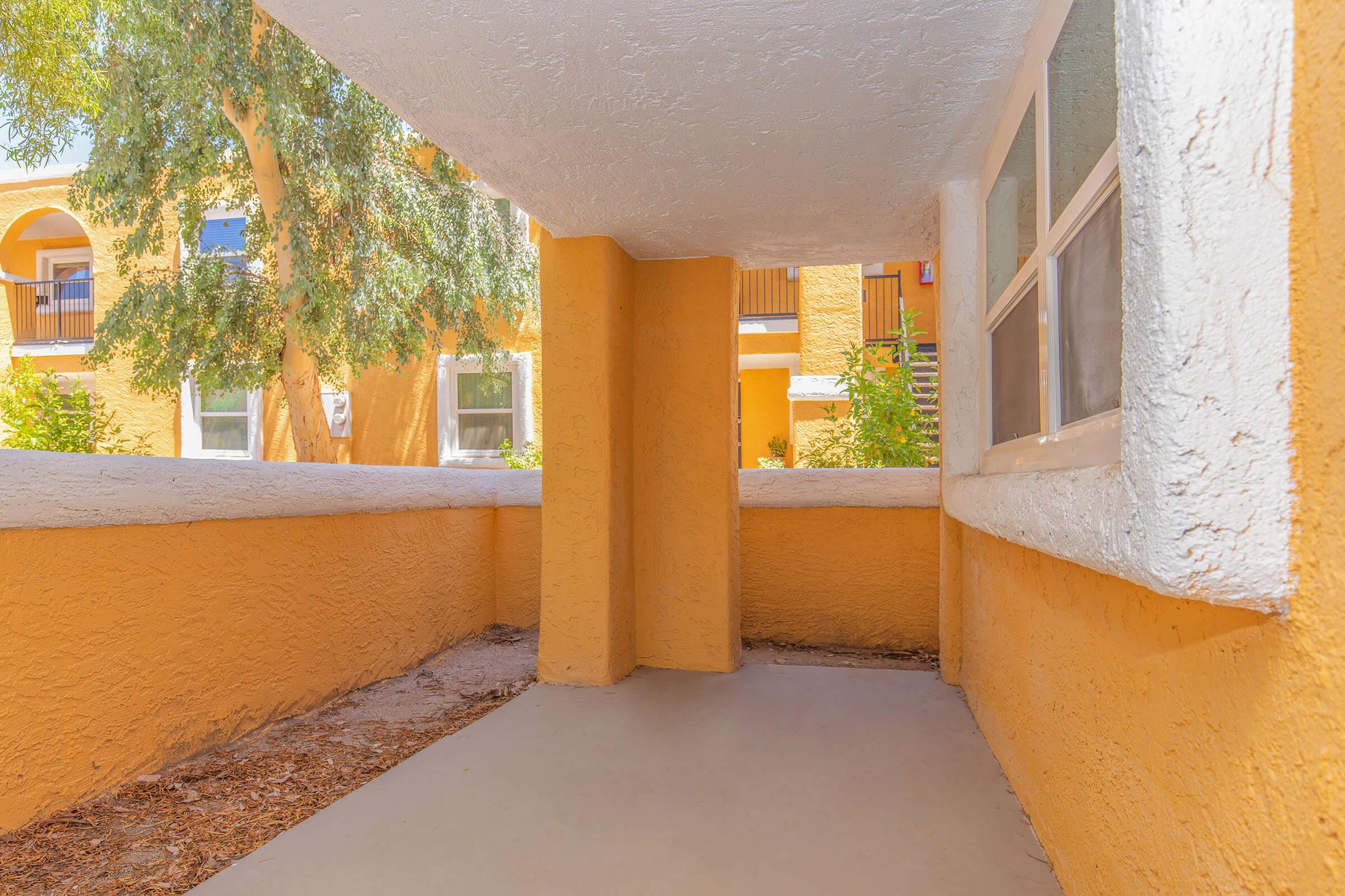 A view of a sunlit corridor featuring textured yellow walls and a clean, empty floor. The corridor leads to an outdoor area with green foliage visible through windows, creating a bright and welcoming atmosphere. Natural light filters in, enhancing the vibrant colors of the architecture.