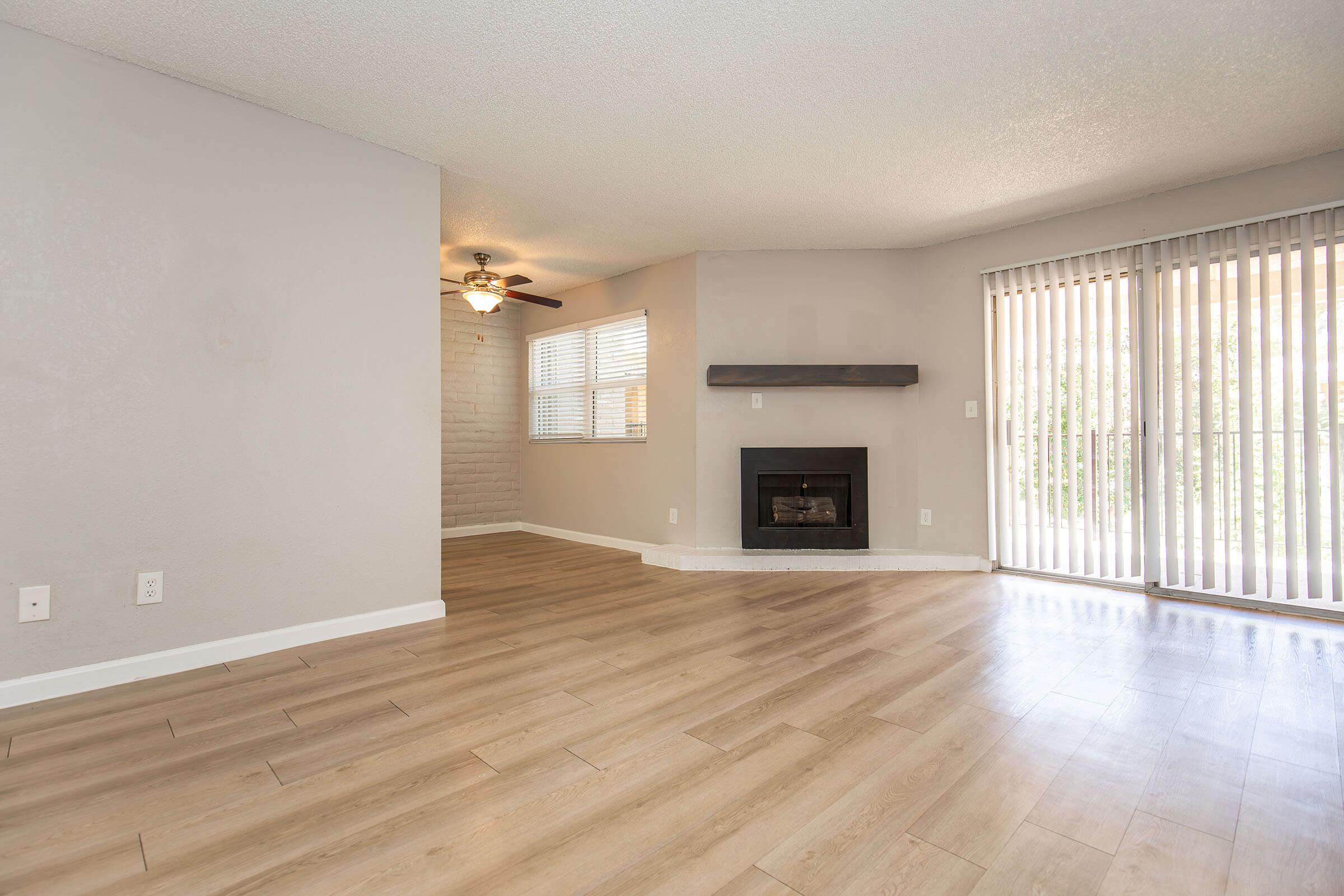 Spacious living area with light brown wooden floors, a modern brick fireplace, and a ceiling fan. Large windows with vertical blinds allow natural light, and the walls are painted in a soft gray tone. A cozy, inviting atmosphere perfect for relaxation or entertaining.