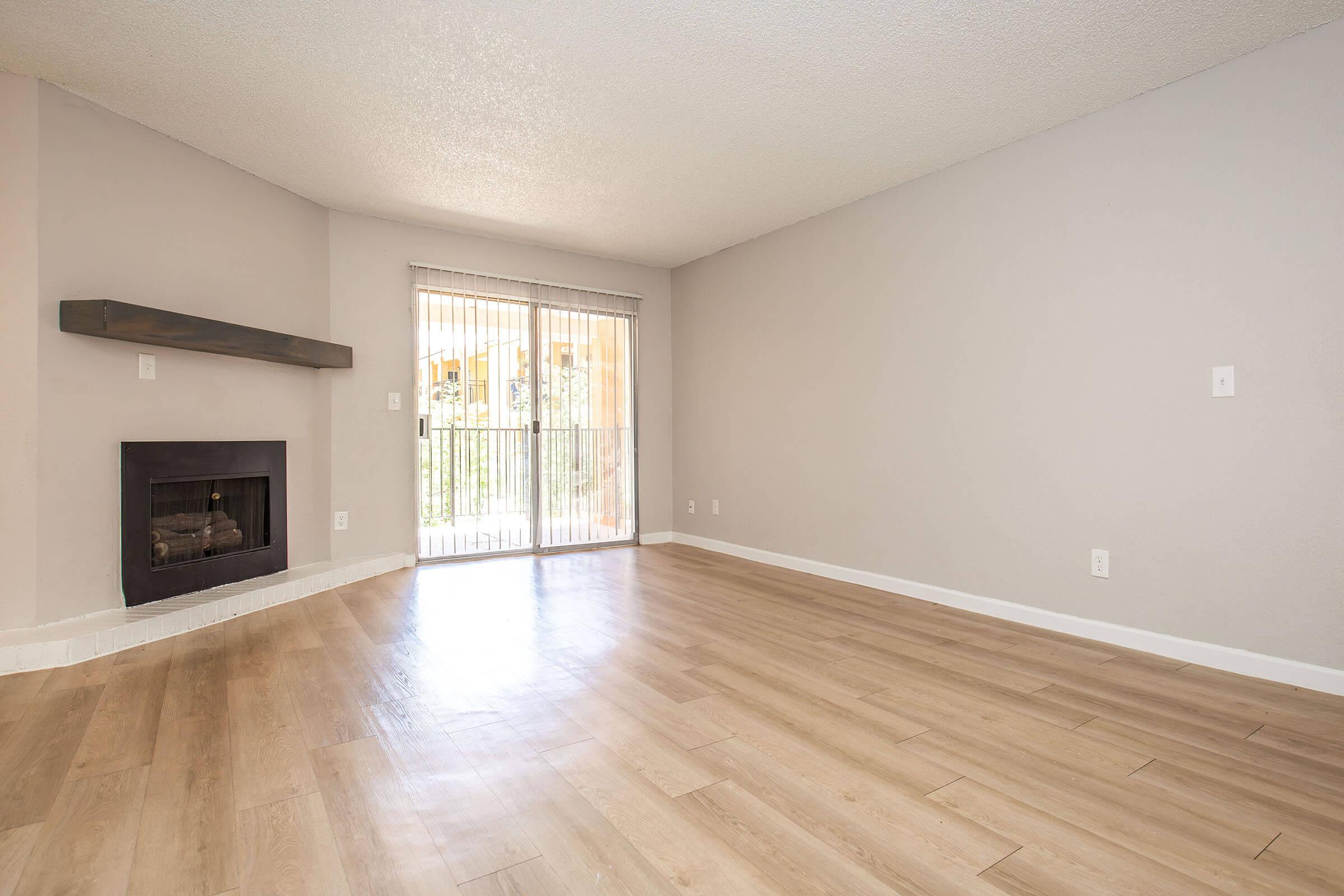 A spacious, empty living room featuring light wood flooring, a modern fireplace on the left, and large sliding glass doors that lead to an outdoor area. The walls are painted in a soft gray, creating a bright and airy atmosphere, with natural light streaming in through the doors.