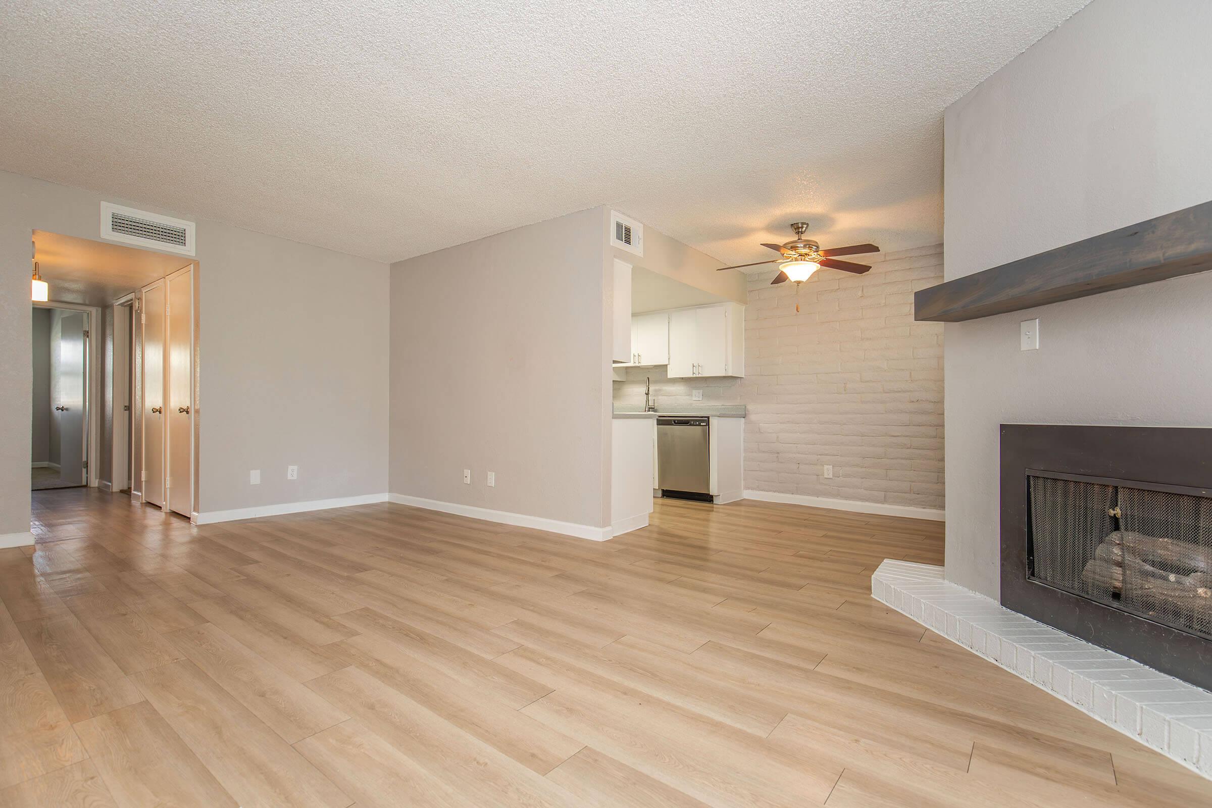 Spacious living area with wood flooring, light gray walls, and a cozy fireplace. The open layout connects to a modern kitchen featuring white cabinetry. A ceiling fan adds comfort, while light streams in from the adjacent room. Clean and inviting space suitable for relaxing or entertaining.