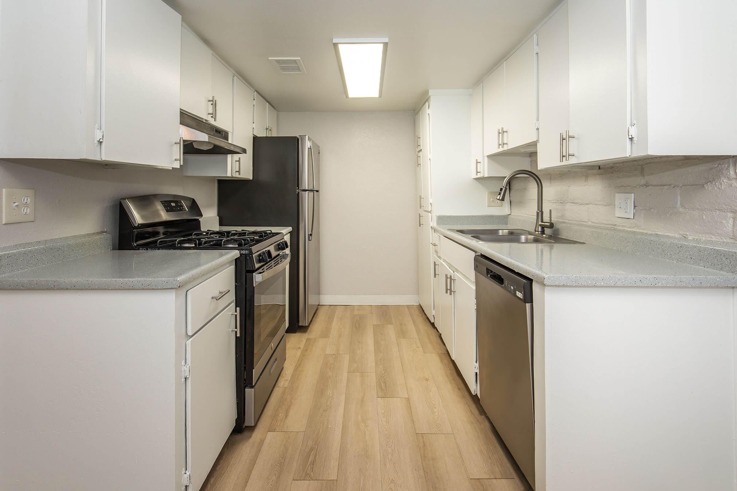 Modern kitchen featuring white cabinetry, stainless steel appliances including a dishwasher, stove, and refrigerator. The countertops are light gray with a subtle texture. The flooring is a warm wood look, and there is overhead lighting providing bright illumination in the space.