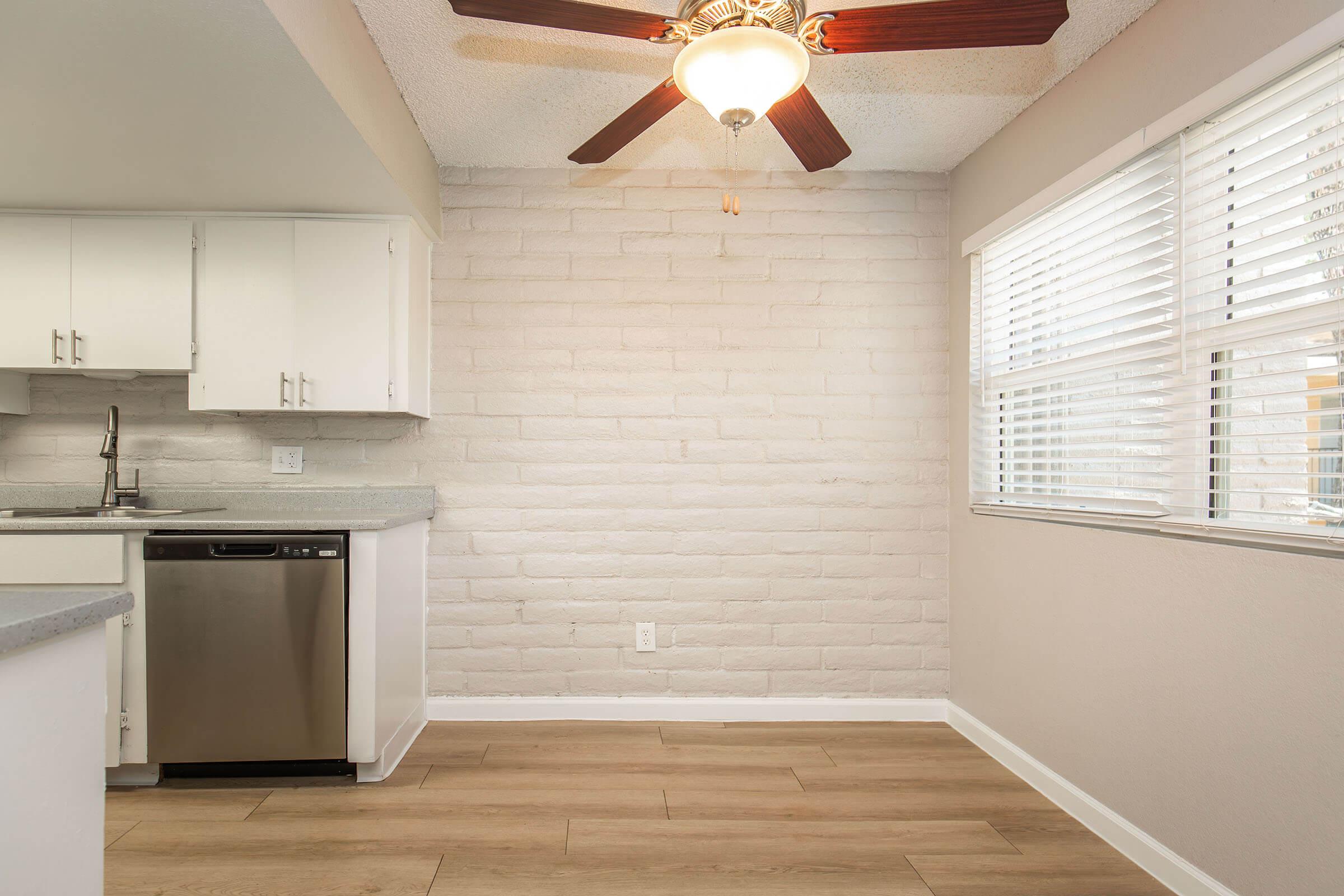 Bright kitchen space featuring a ceiling fan, light fixture, and white brick wall. The area includes a countertop, stainless steel dishwasher, and several cabinets. Natural light streams in through the window with white blinds, illuminating the warm wood-like flooring.