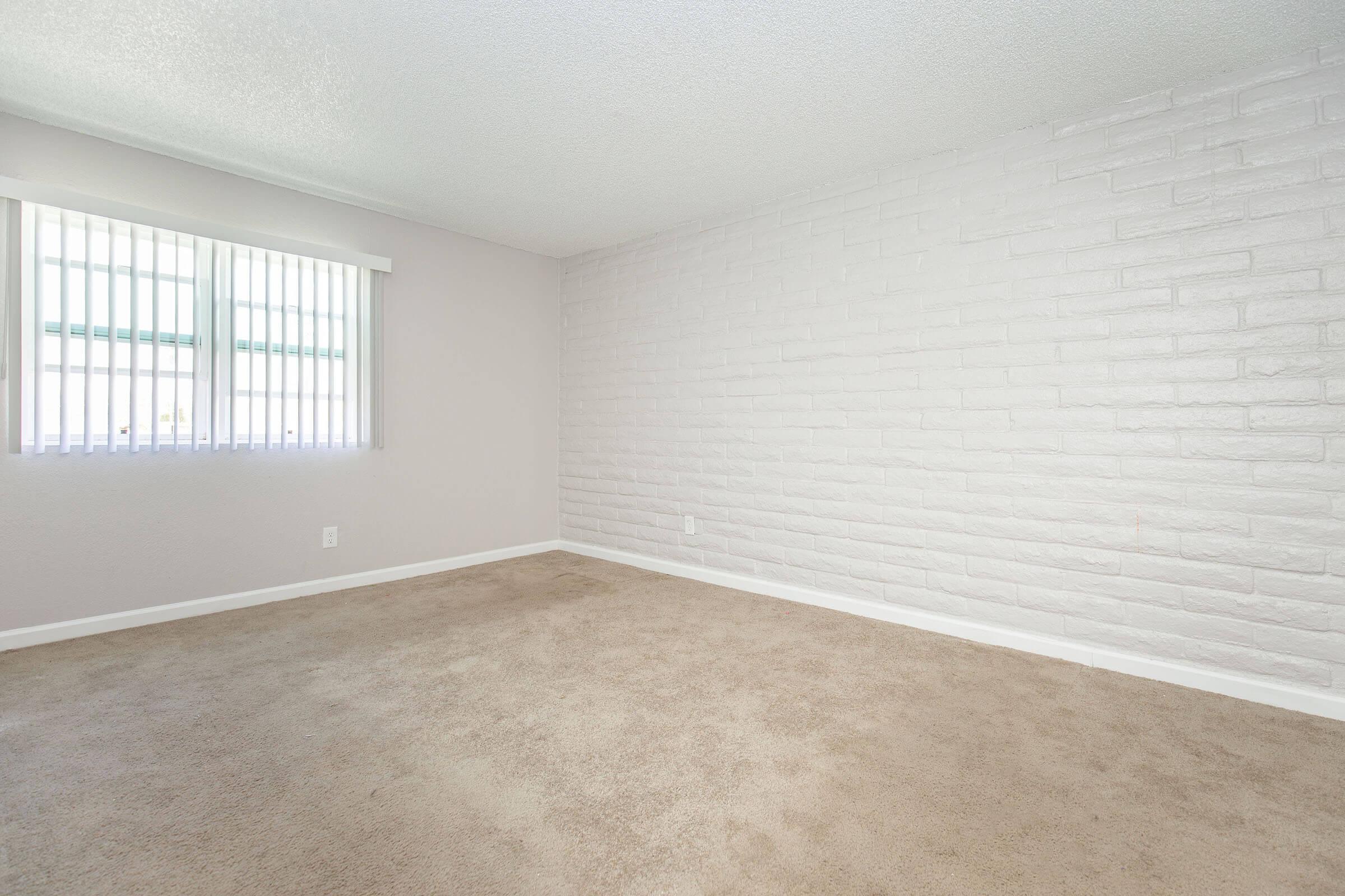 A spacious, empty room with light beige carpet. One wall features textured white bricks, while another has two narrow windows with vertical blinds. The overall atmosphere is bright and inviting, suitable for various furnishing options or personal decoration.