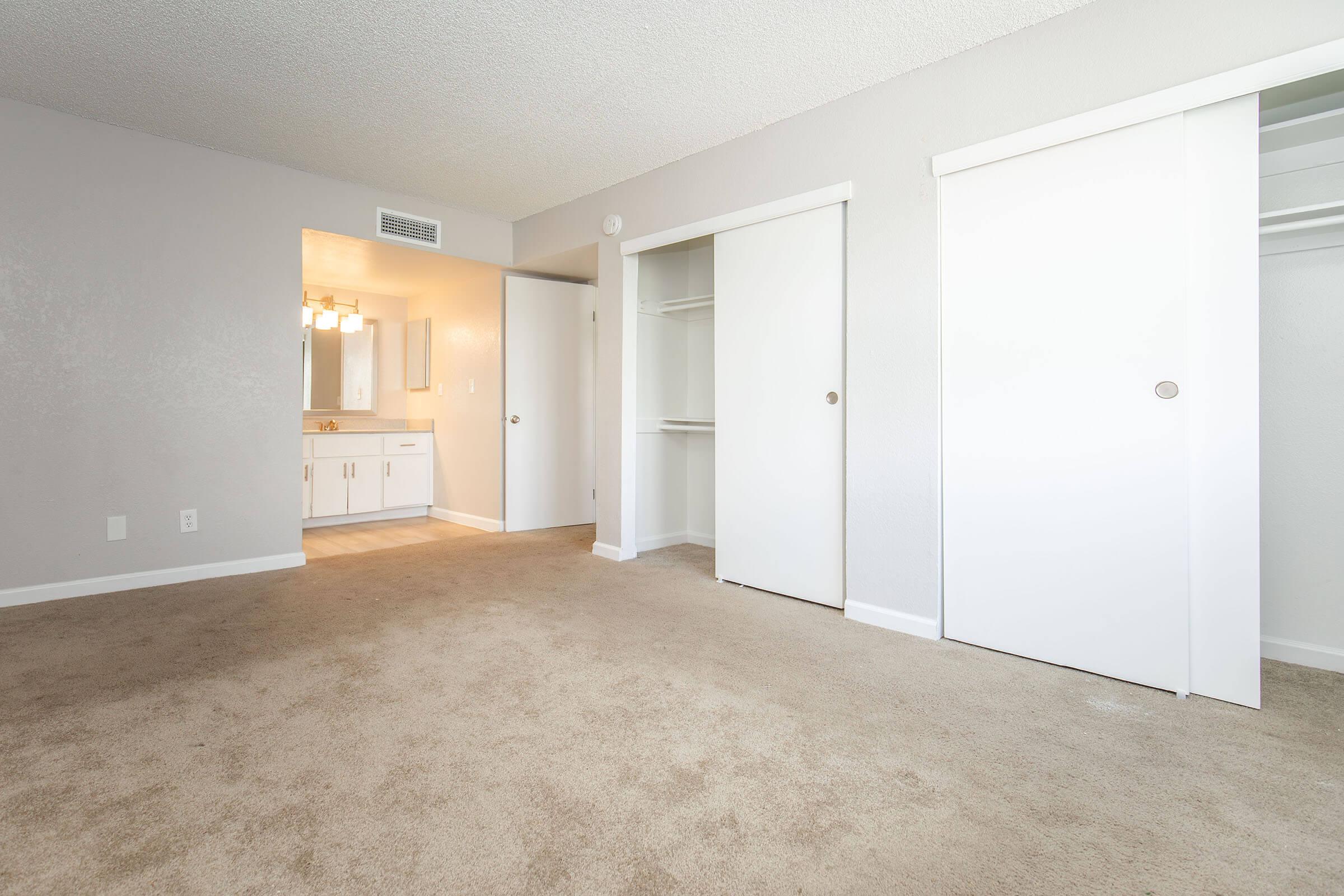 A spacious, neutral-colored bedroom featuring beige carpet. Two white sliding closet doors are visible, along with a doorway leading to a bathroom area with vanity lighting. The walls have a light gray finish, and natural light fills the room, creating a bright and airy atmosphere.