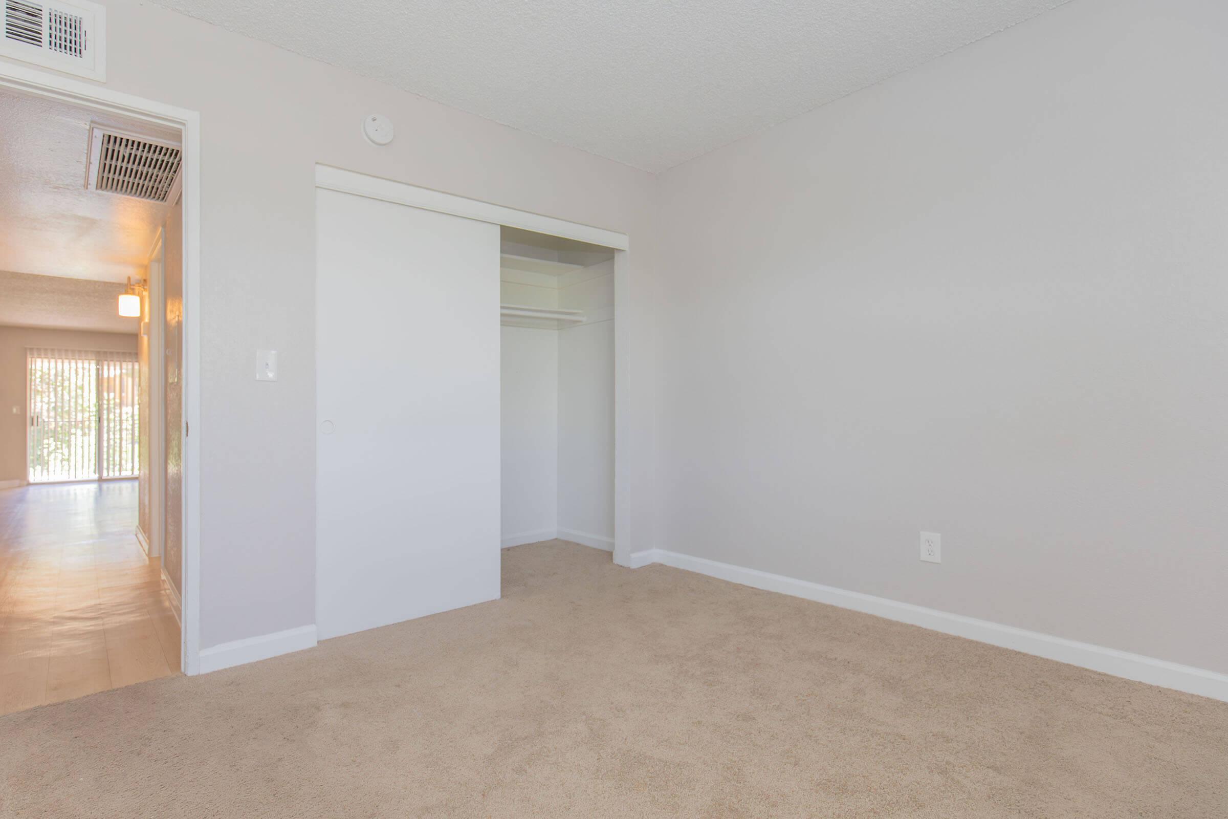 A clean, empty room with light beige carpeting and light gray walls. There is a closet with sliding doors on one side and a doorway leading to another space in the background, suggesting a well-lit and open layout. The overall appearance is modern and inviting.