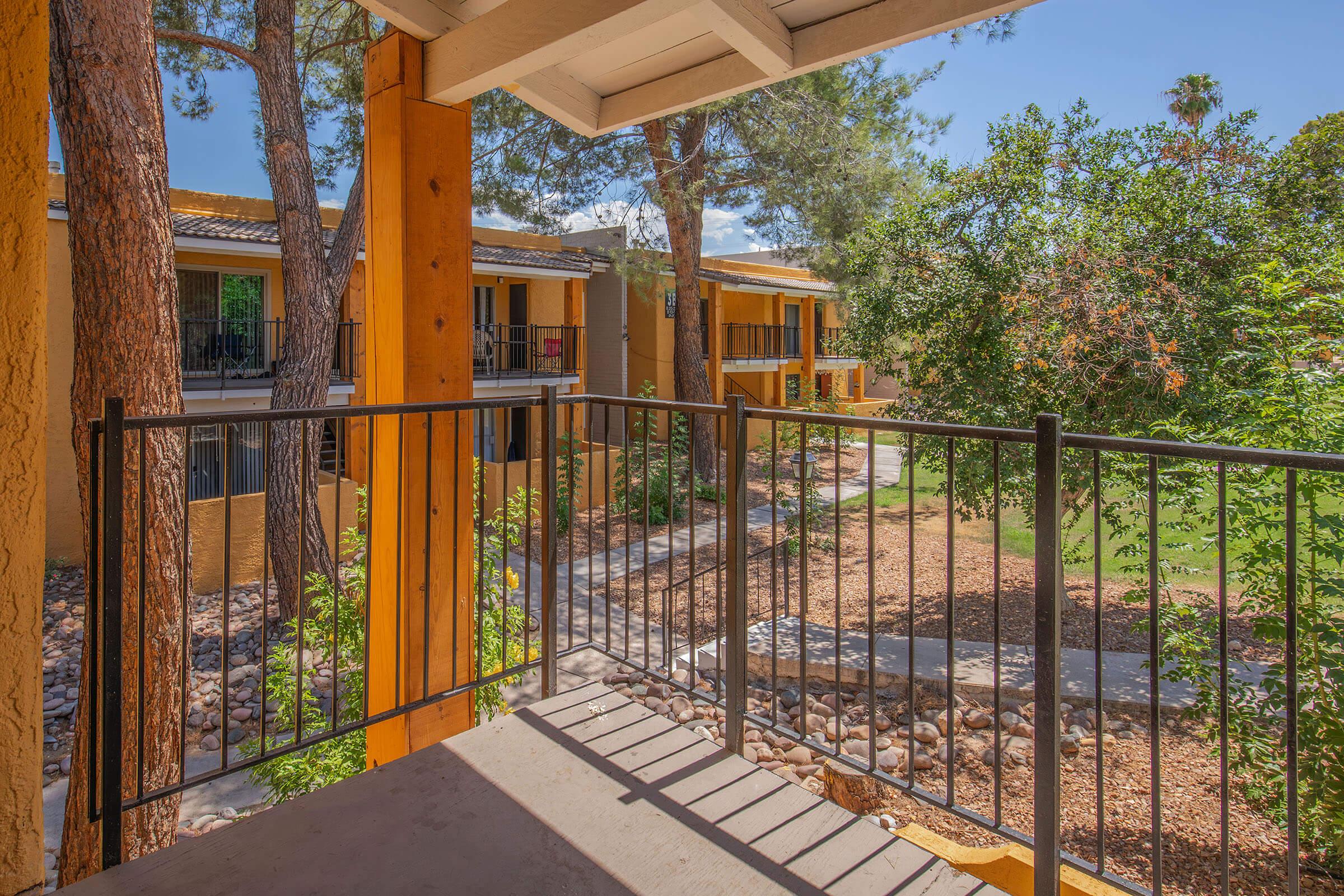 View from a balcony overlooking a landscaped courtyard with trees, shrubs, and a pathway connecting adjacent buildings. The scene features a sunny sky and a peaceful atmosphere, highlighting the architectural details of the railing and the surrounding greenery.