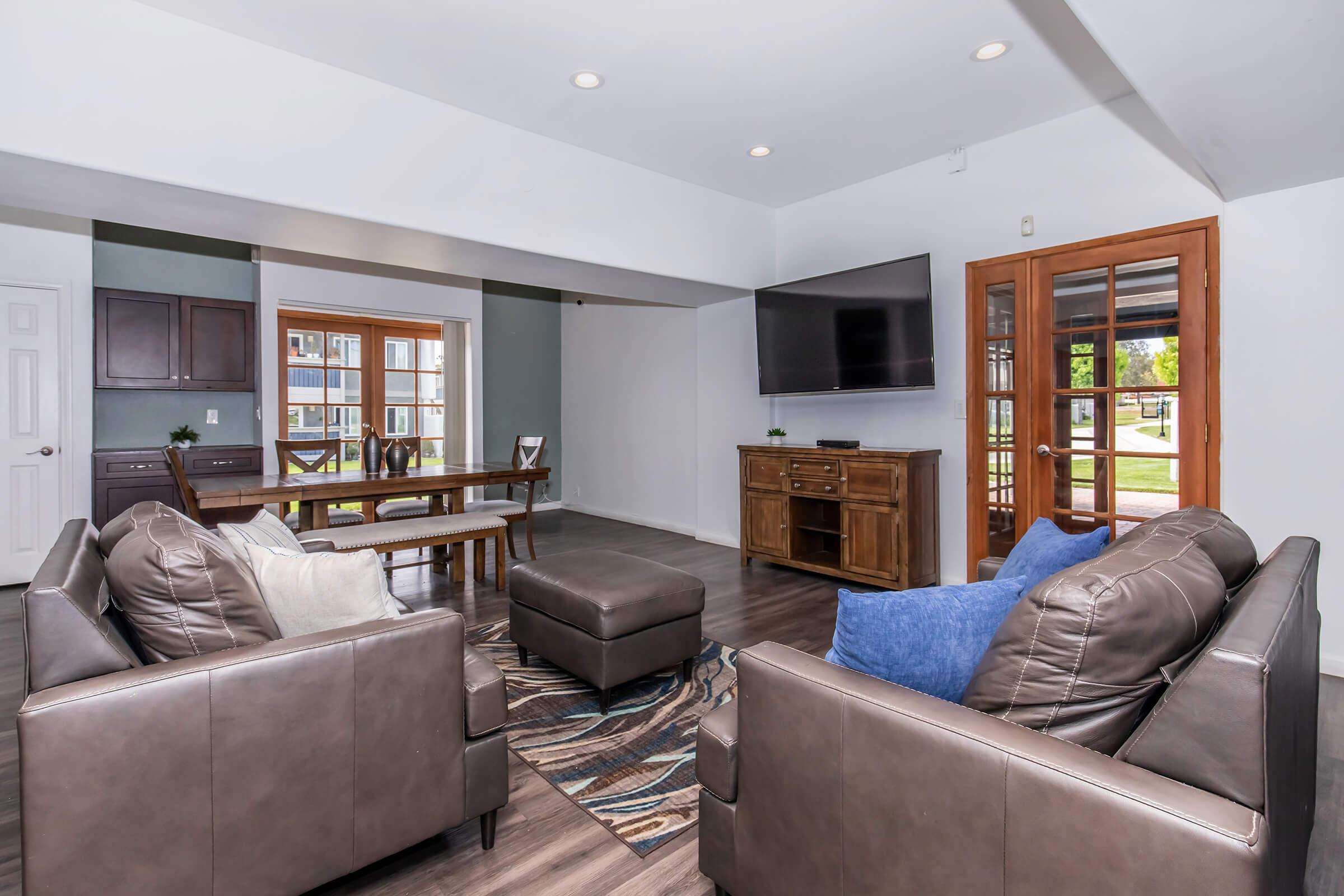 A modern living room featuring two brown leather chairs and a matching ottoman. In the background, there is a wooden dining table with chairs, a TV mounted on the wall, and large windows showing outdoor views. The floor is covered with a decorative area rug.