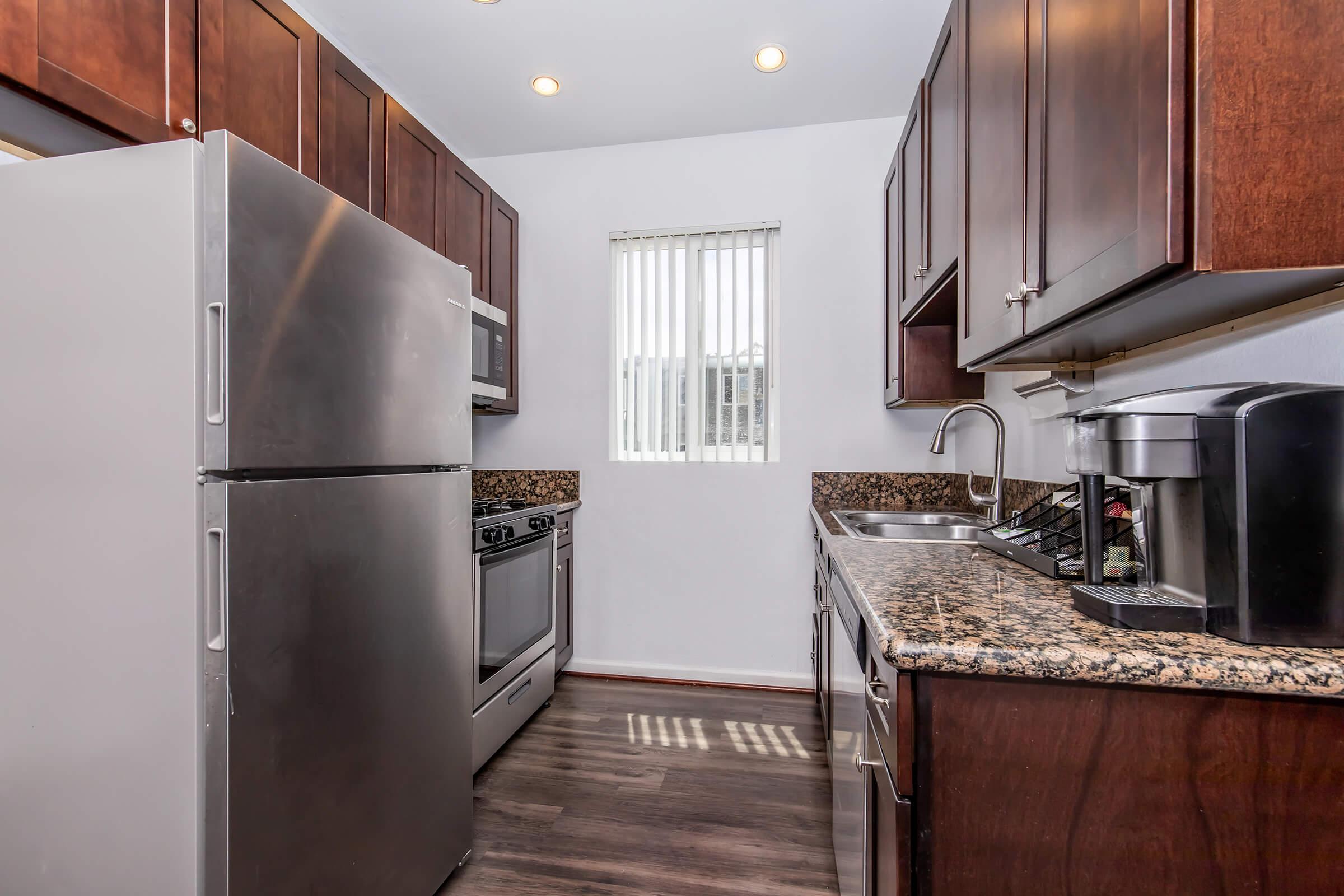 A modern kitchen featuring dark wooden cabinets, stainless steel appliances including a refrigerator, oven, and microwave. The countertops are made of granite. A small window with vertical blinds allows natural light to enter the space, and a coffee maker is placed on the counter. The flooring is dark and the overall design is contemporary.