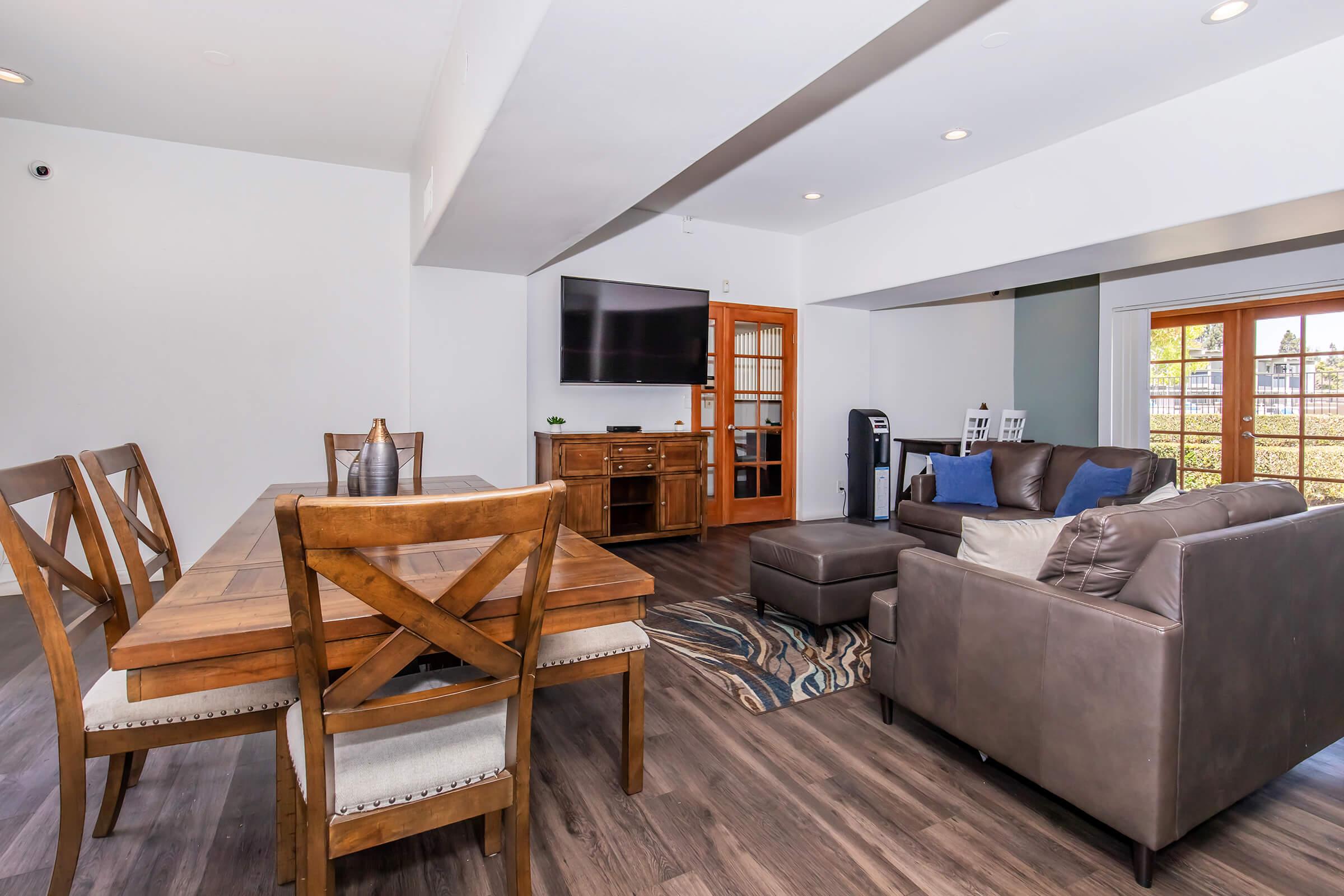 An inviting modern living space featuring a wooden dining table with chairs, a comfortable brown sofa, an ottoman, and a TV mounted on the wall. Large windows allow natural light, and there are decorative elements like a rug and plants, creating a warm and cozy atmosphere.