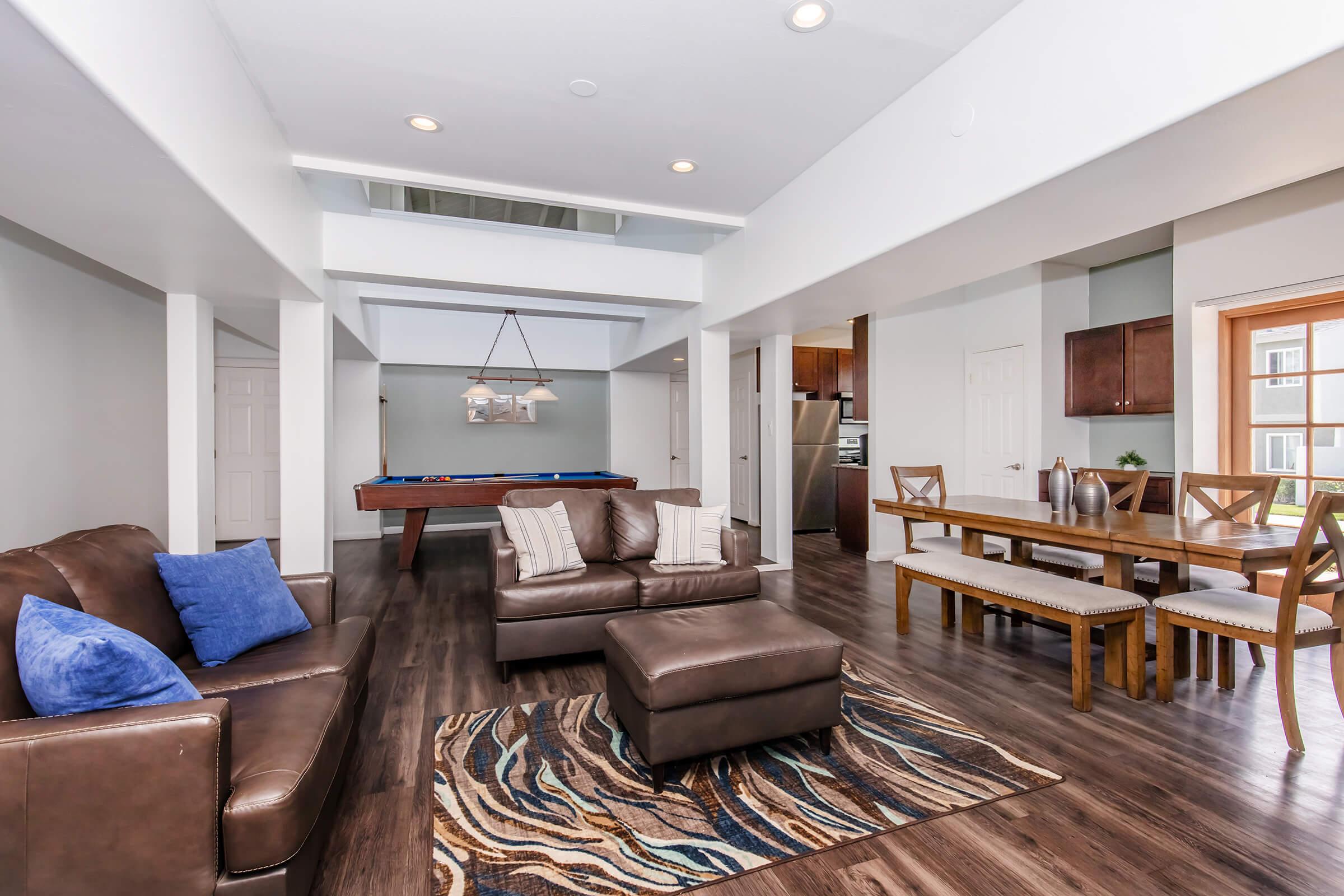 A spacious living room featuring a brown leather sofa with blue cushions, a patterned area rug, and a coffee table. An adjacent dining area has a wood table with chairs, and a kitchen area visible in the background. Natural light fills the space, highlighting modern decor and an inviting atmosphere.