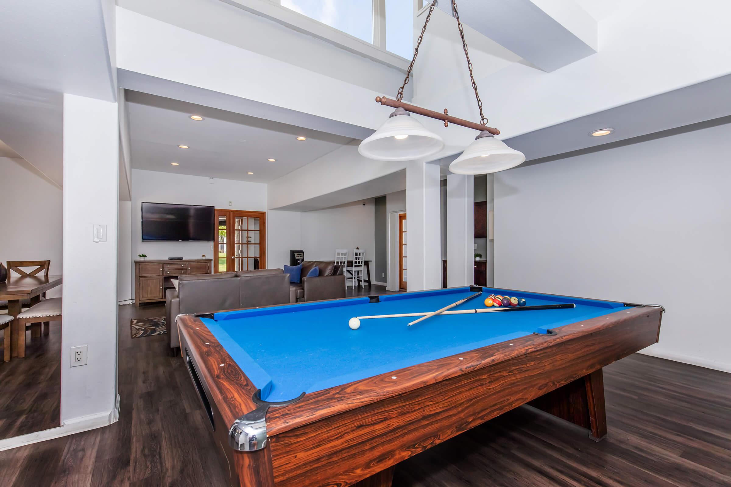 Interior shot of a spacious game room featuring a blue felt pool table with a cue and balls. The room is well-lit with recessed lighting and a large window, and there are modern furnishings including a sofa and a television in the background. The flooring is dark wood, creating a warm atmosphere.