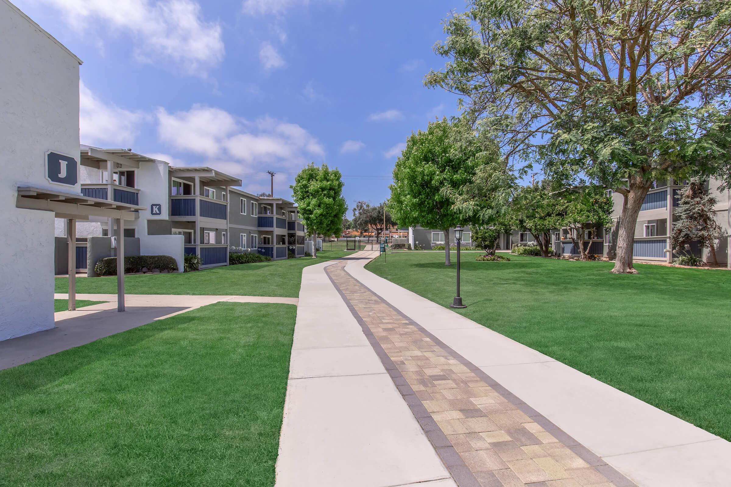 A paved walkway meanders through a well-maintained green space, flanked by trees and residential buildings. The sky is partly cloudy, creating a serene atmosphere in a community setting.