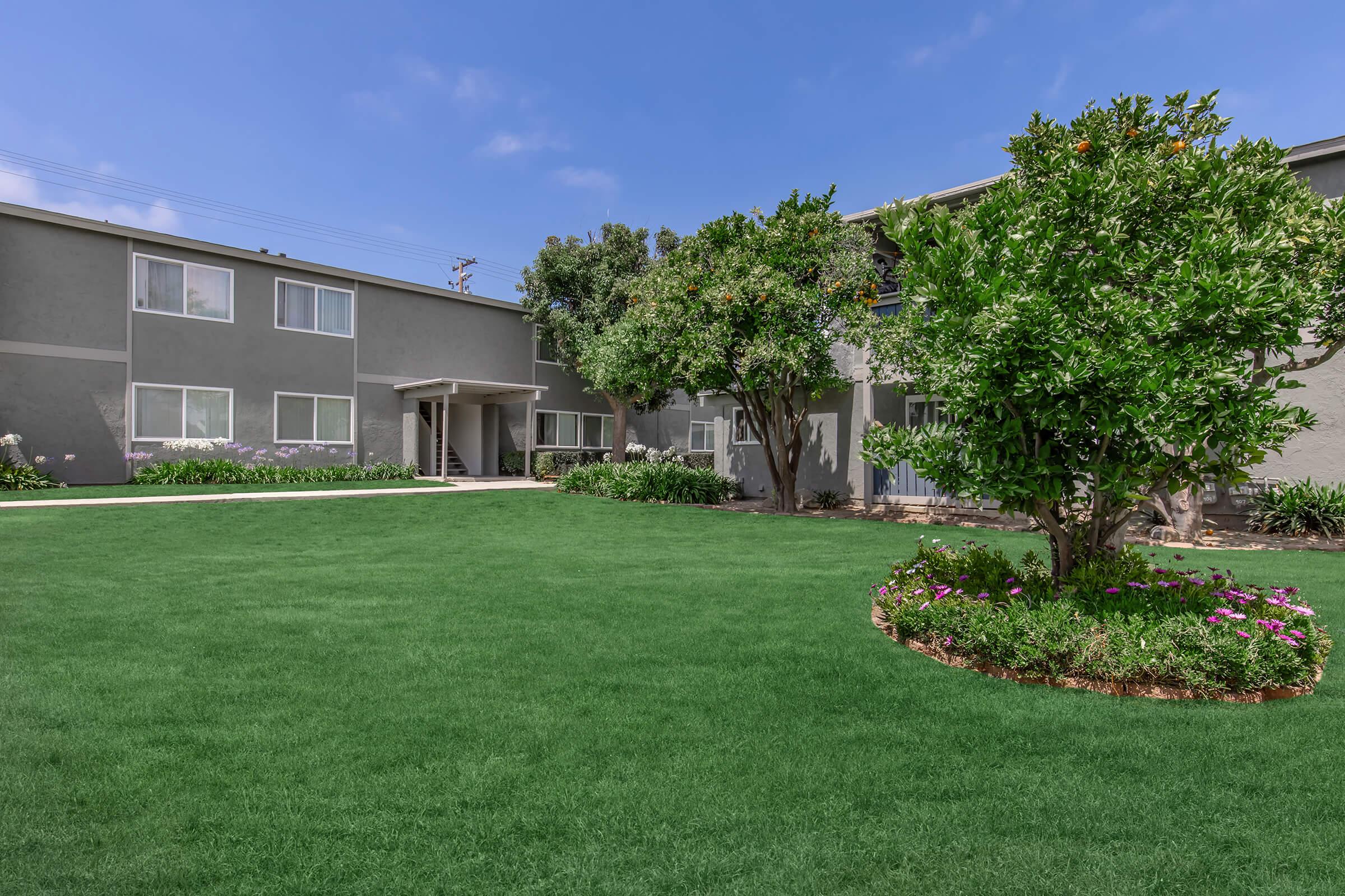 A well-maintained outdoor area featuring a vibrant green lawn, surrounded by landscaped flower beds. Two gray apartment buildings are visible in the background, with trees providing shade and adding to the peaceful ambiance of the space. The sky is clear with some clouds.