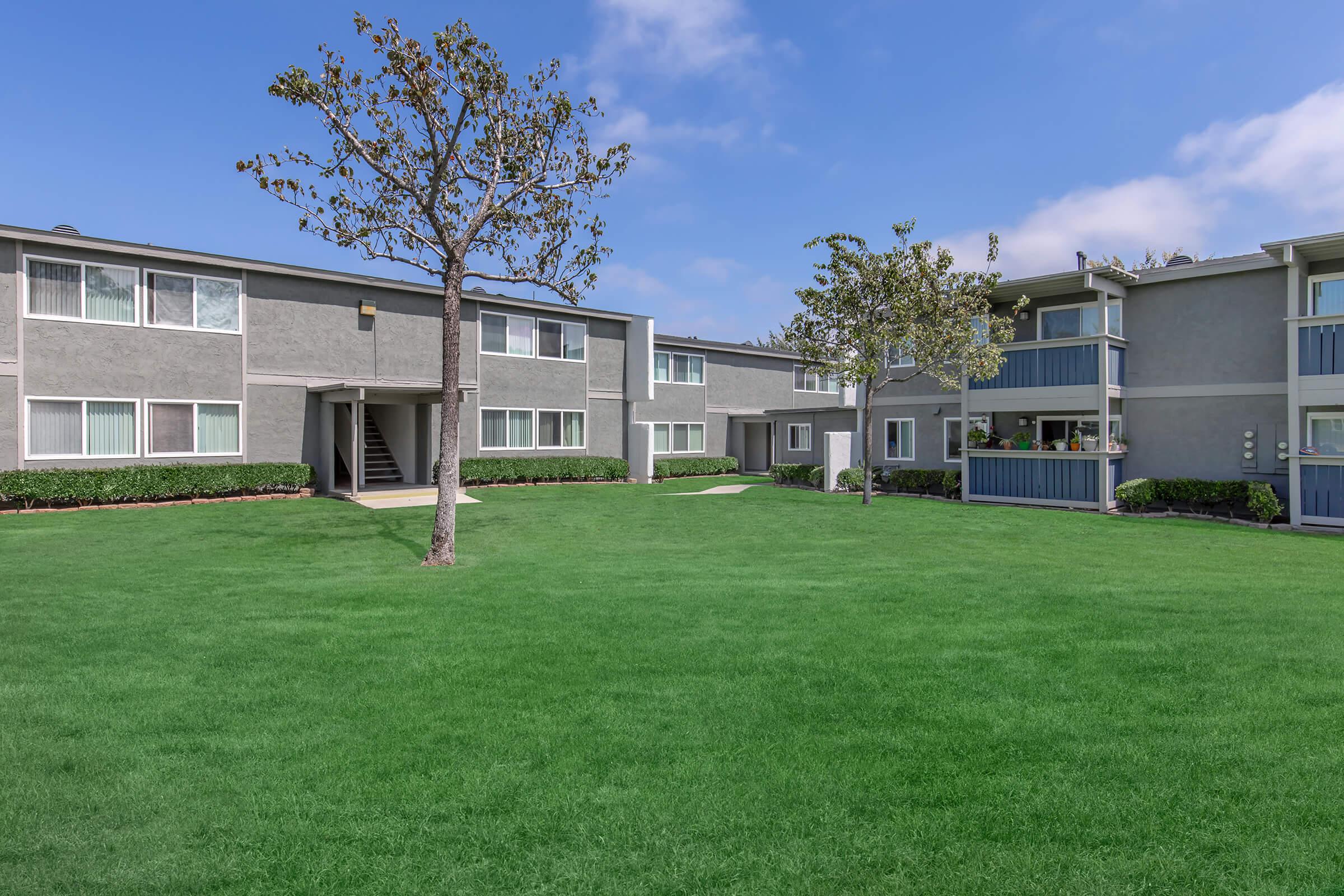 Lush green lawn in a landscaped apartment complex, featuring two-story buildings with gray exteriors. A small tree stands prominently in the foreground, and balconies can be seen on some of the upper floors. The sky is clear with a few scattered clouds.