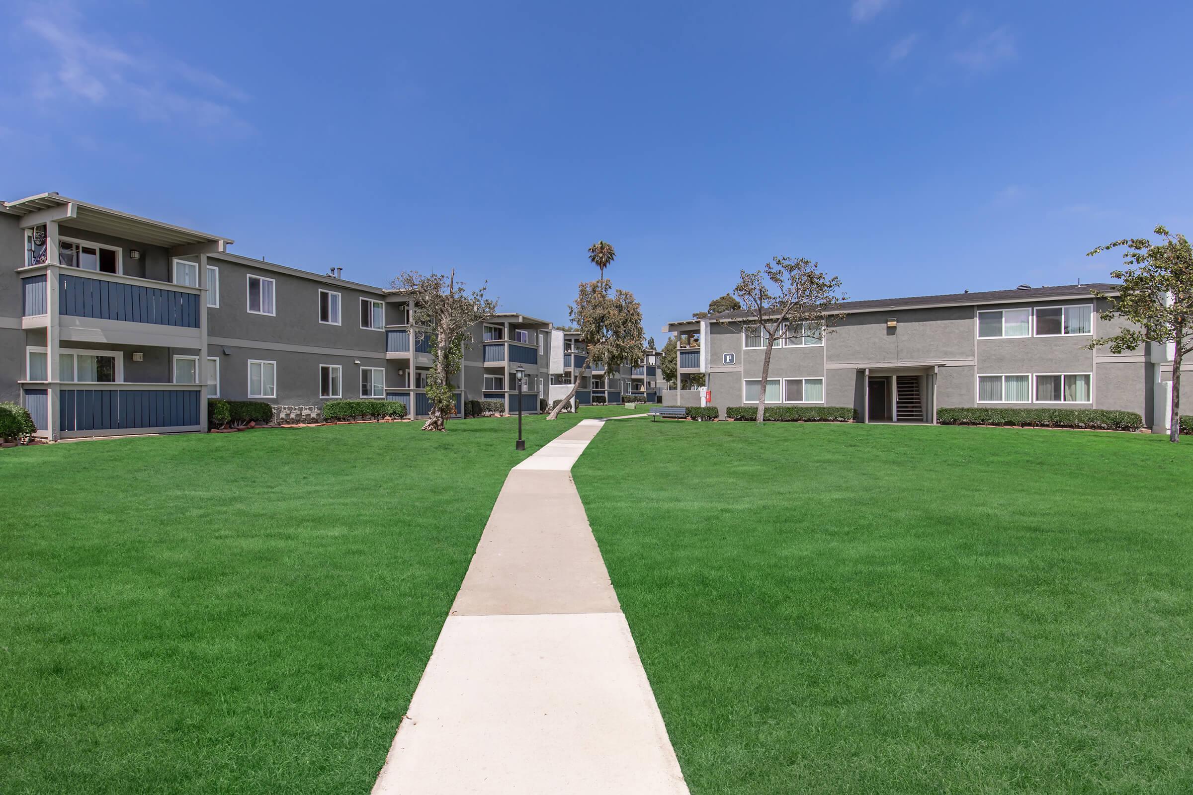 A well-kept residential complex featuring two-story apartment buildings arranged around a green lawn. A concrete pathway runs through the center, leading to the buildings. The sky is clear with a few clouds, and there are palm trees and landscaping around the area, creating a peaceful suburban environment.
