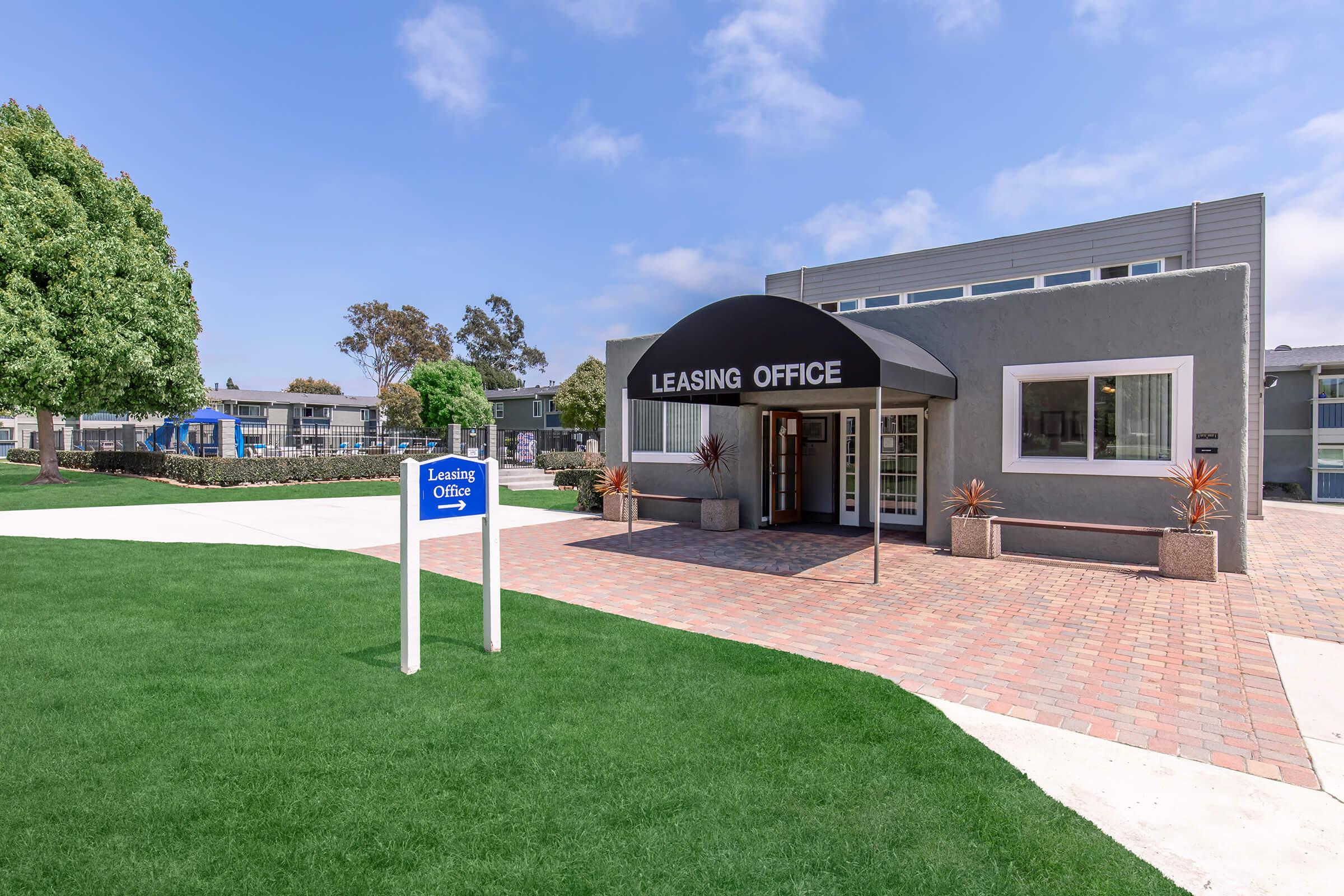 Leasing office building with a black awning and sign, surrounded by green grass and trees. A blue sign with "Leasing Office" is visible in front. The entrance features large windows, and the area includes a paved walkway leading to the building. Clear blue sky in the background.