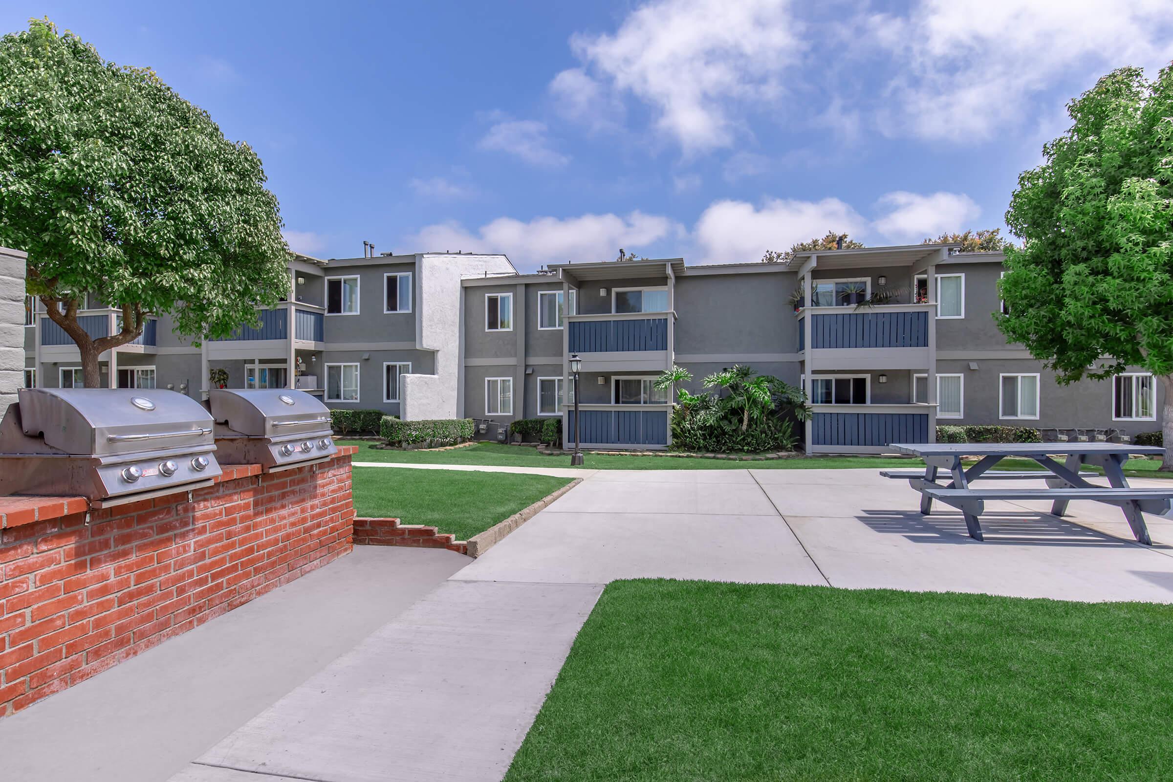 A well-maintained apartment complex with multiple buildings. In the foreground, there are barbecue grills and a picnic table on a grassy area. The background features the apartment units, which have balconies and windows, under a clear blue sky with some clouds.