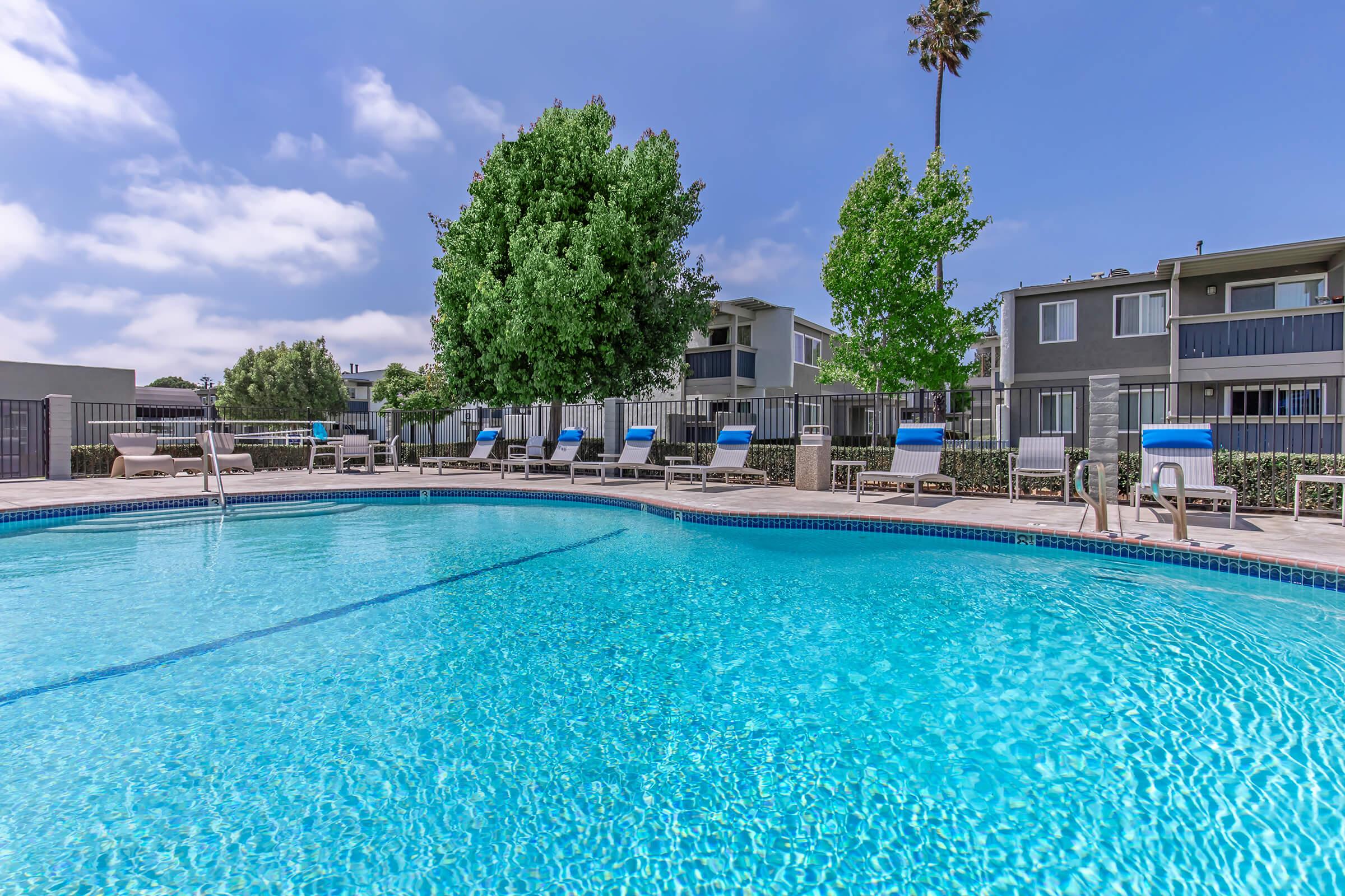 A clear blue swimming pool surrounded by lounge chairs, with green trees and modern apartment buildings in the background under a partly cloudy sky.