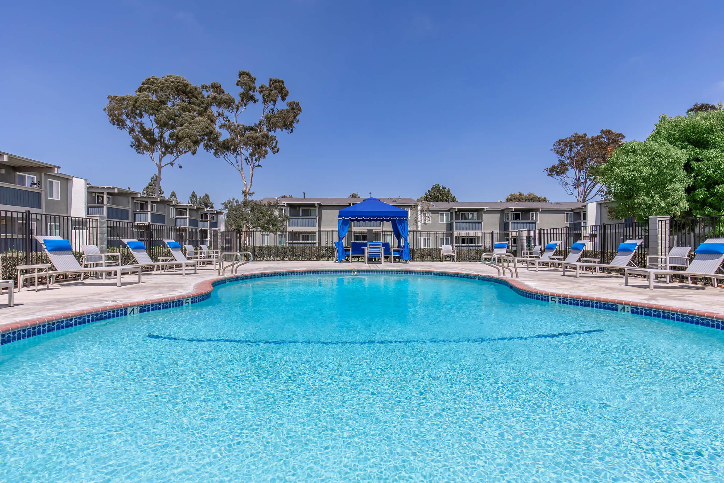 A large swimming pool with clear blue water, surrounded by lounge chairs. In the background, several apartment buildings are visible. A blue gazebo offers shade near the poolside, with trees lining the area, creating a relaxing atmosphere under a bright blue sky.