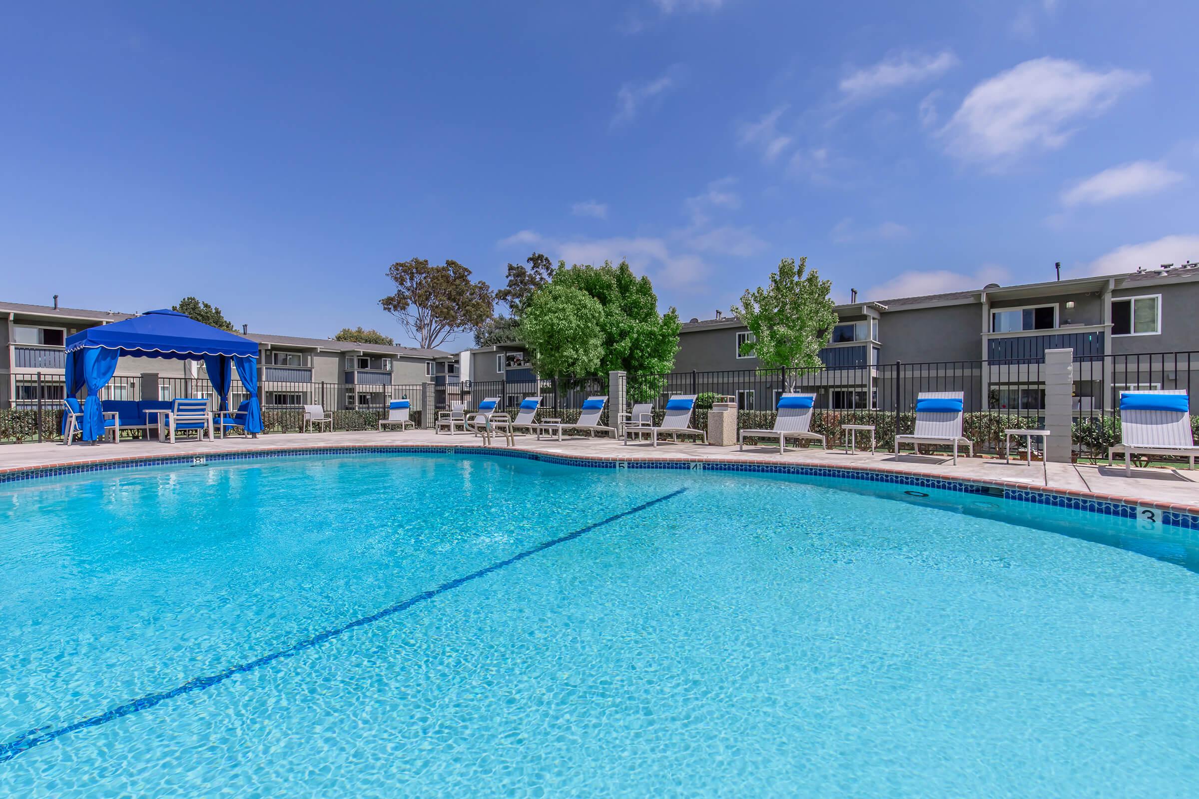 A sunlit swimming pool surrounded by lounge chairs, with a blue canopy on one side. The pool reflects the clear blue sky and is set against a backdrop of landscaped trees and residential buildings. Ideal for relaxation and leisure activities.