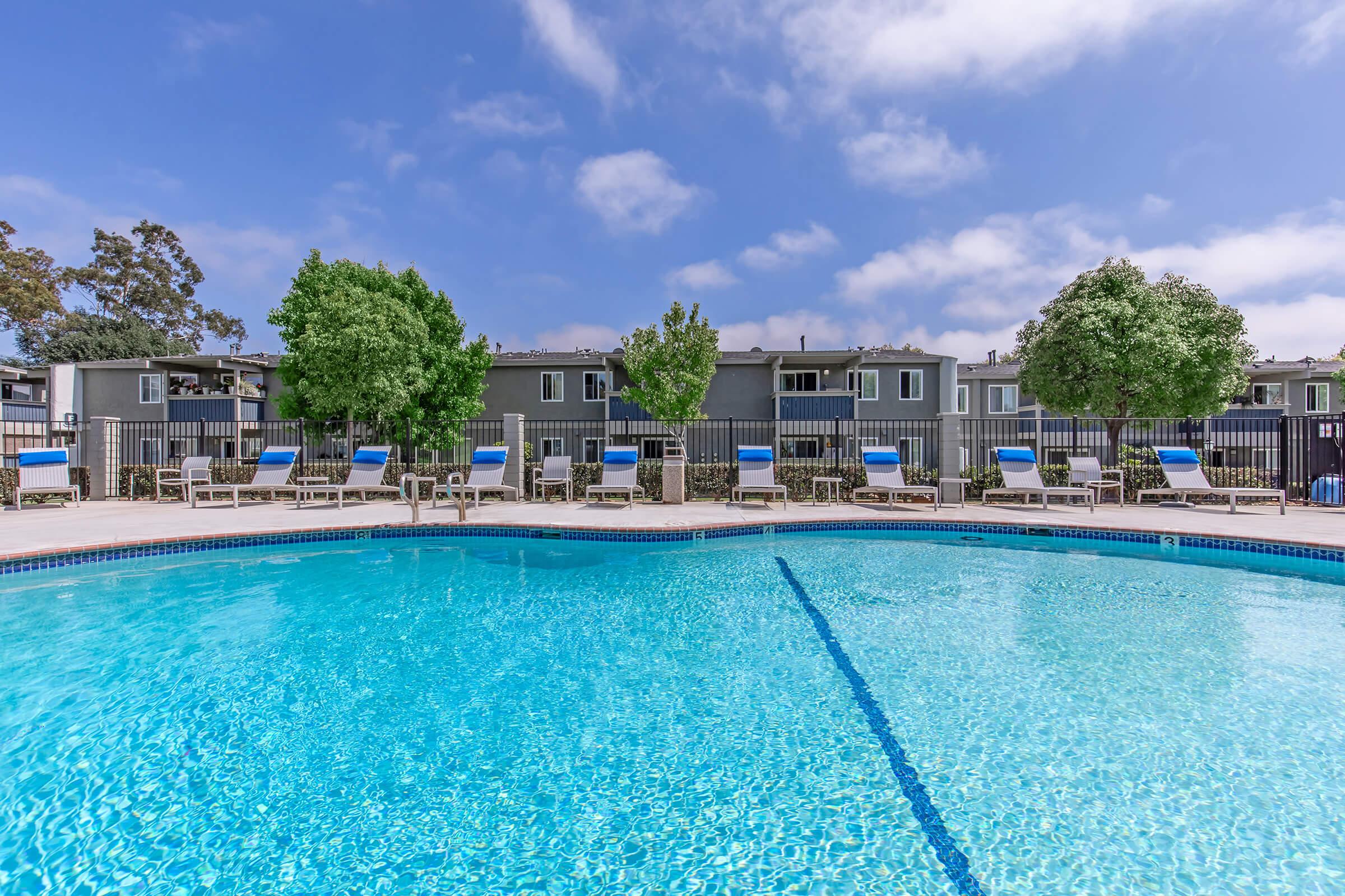 A clear blue swimming pool surrounded by lounge chairs and trees. In the background, there are residential buildings enclosed by a fence, under a bright blue sky with some clouds.