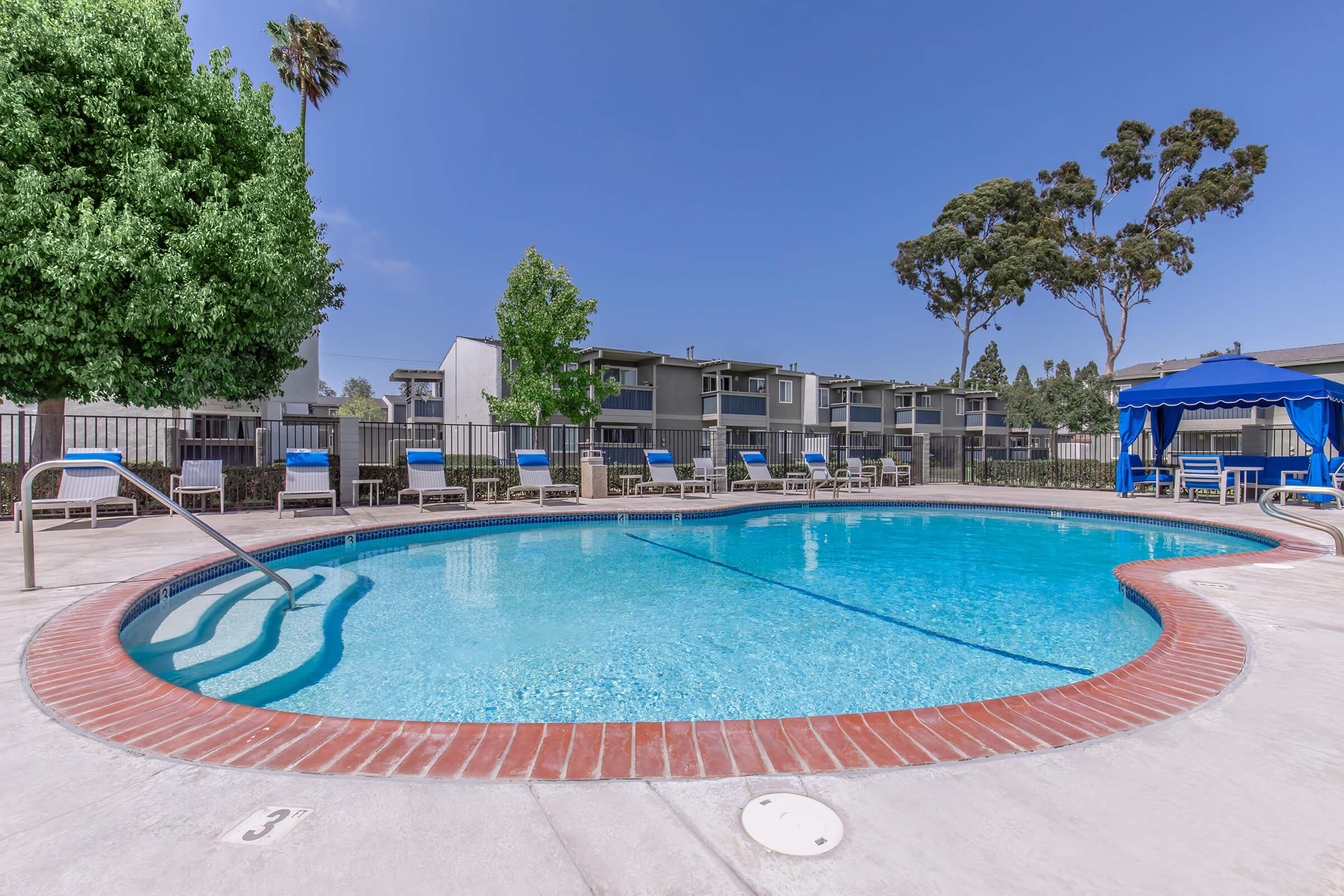 A clear swimming pool surrounded by lounge chairs under a sunny sky. Green trees are visible in the background, along with apartment buildings. A blue cabana is located near the pool, creating a relaxing outdoor space.