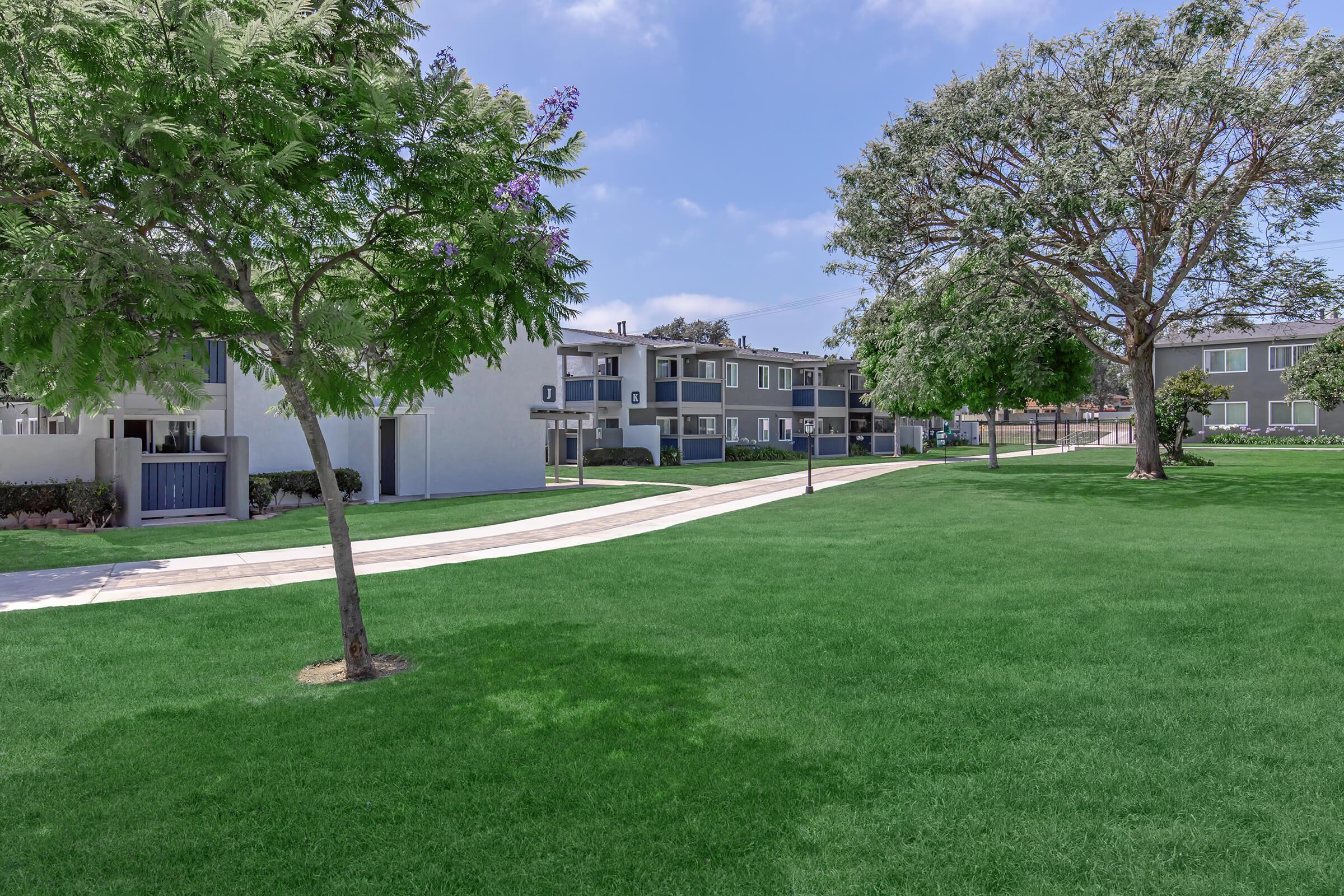 A landscaped area featuring a well-maintained grassy lawn with trees. In the background, there are modern apartment buildings and a paved walkway, creating a serene and inviting residential environment. The sky is clear with a few clouds, enhancing the peaceful atmosphere.
