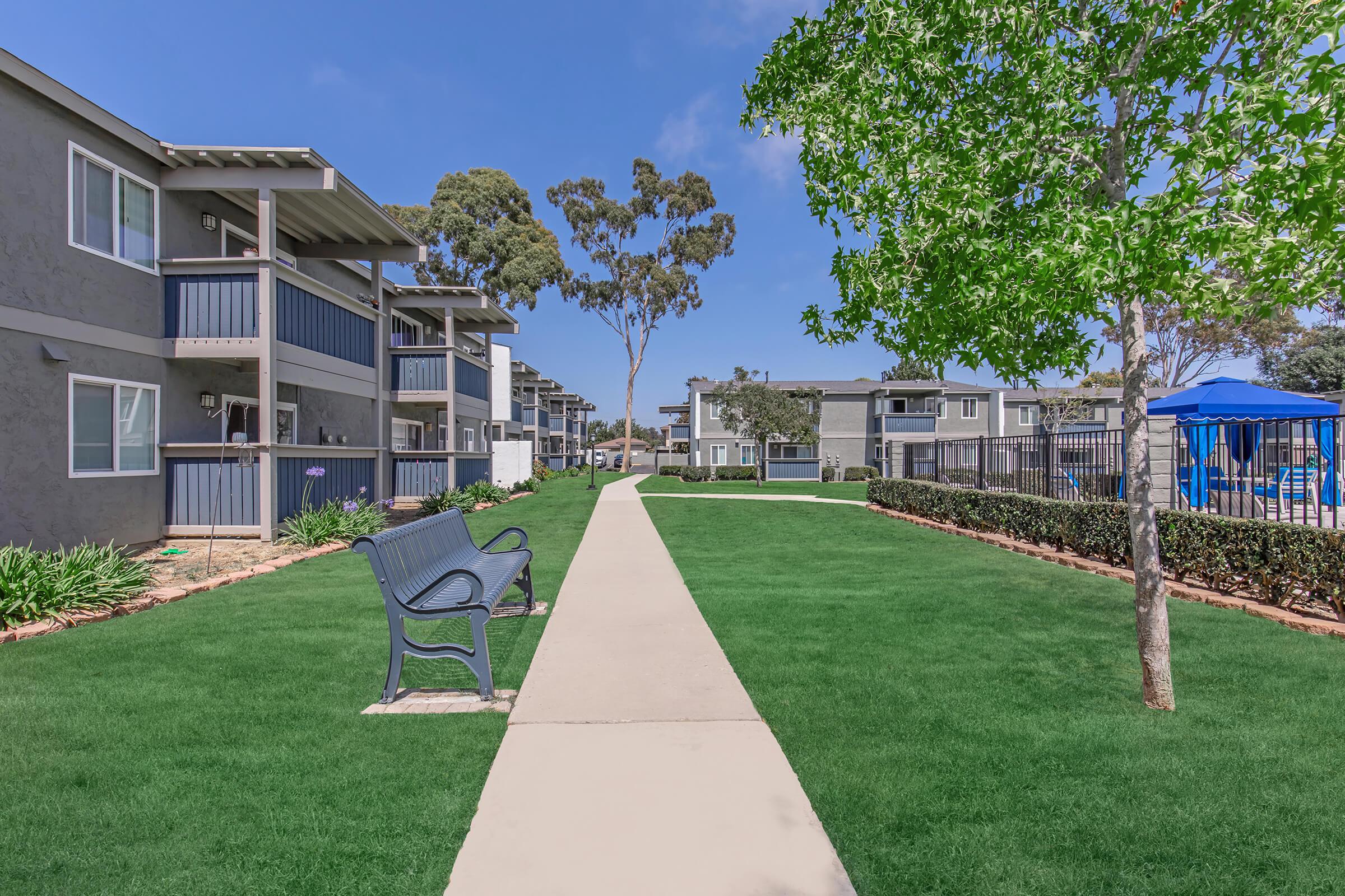A neat pathway lined with green grass leads between modern apartment buildings. Benches are placed along the path, and trees provide shade. In the background, there's a blue pool area, surrounded by fencing and more greenery, creating a welcoming community atmosphere.